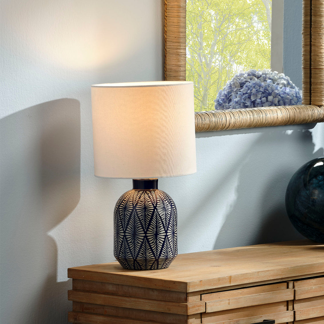 Table lamp with decorative base on a wooden surface, with a mirror and plant in the background.