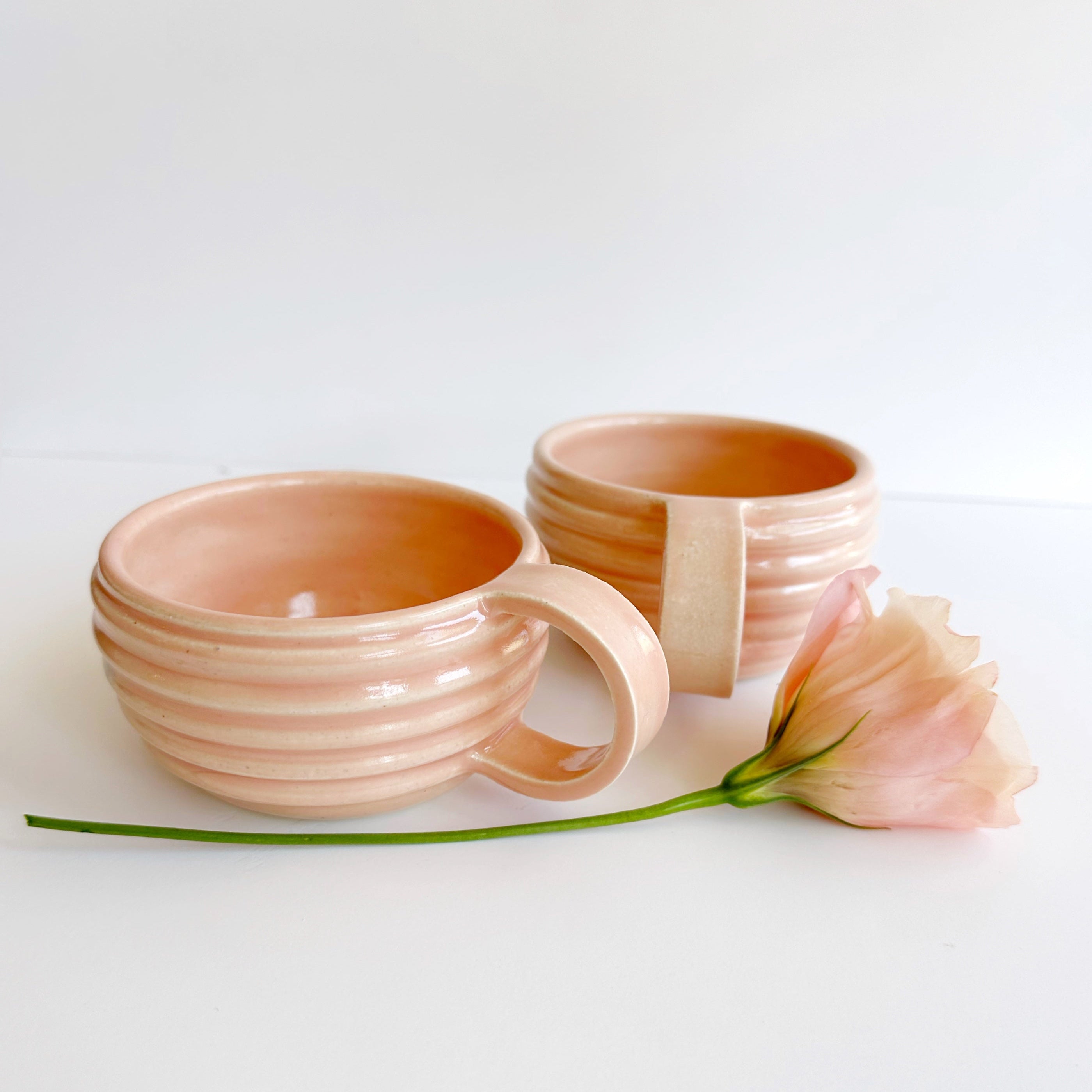 Two pink ceramic mugs on a white surface with a single pink flower.