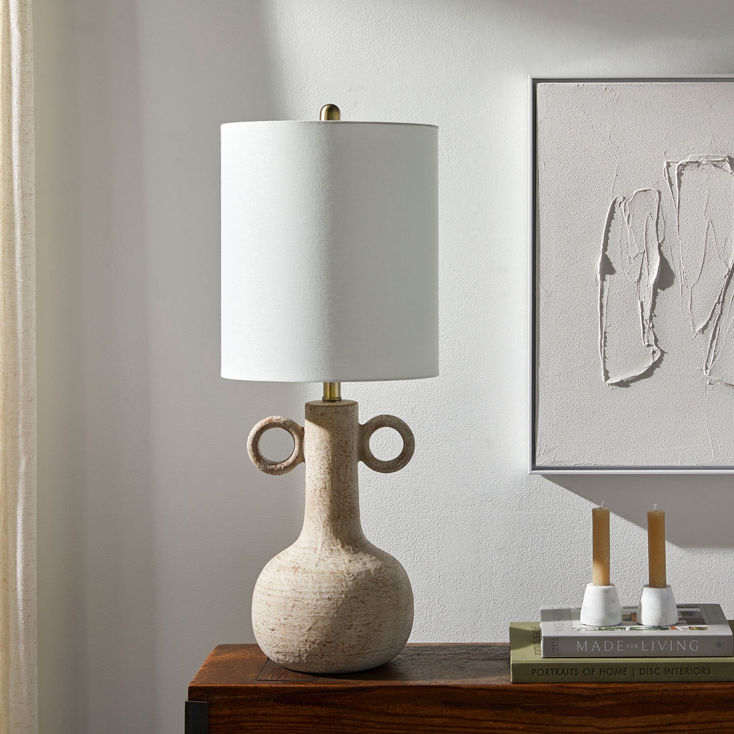 Table lamp with a textured base on a wooden surface, with a neutral wall and abstract art in the background.