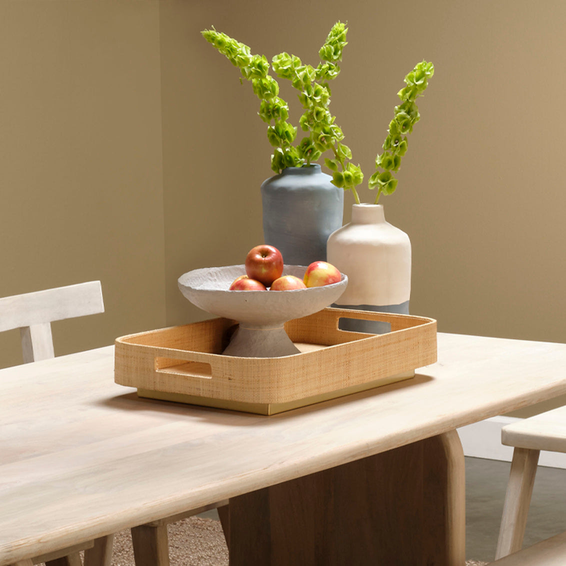 Bamboo tray with fruit and vases on a wooden table against a plain wall.