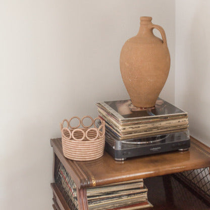 Terracotta vase on a stack of records with a woven basket on a wooden surface against a plain wall.