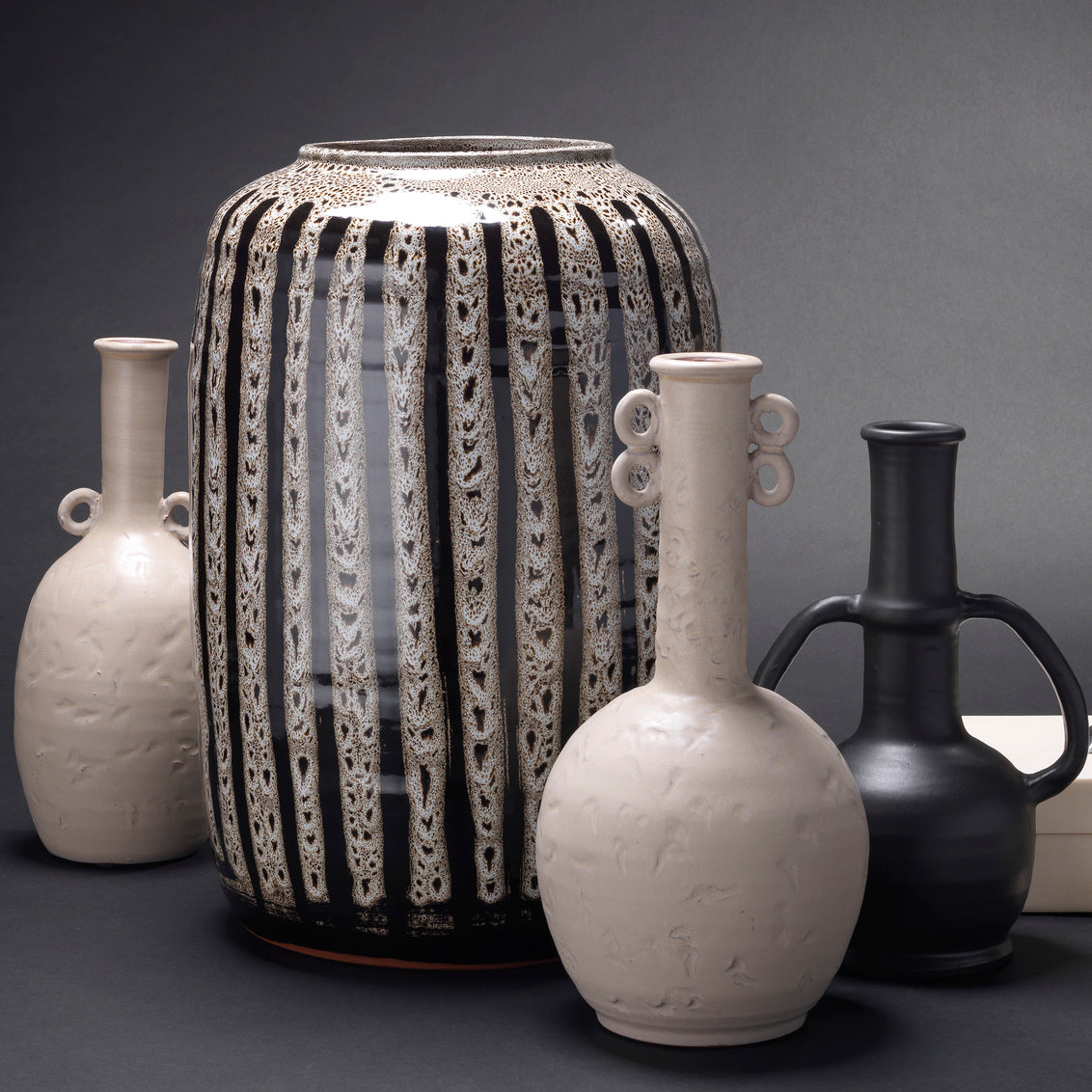 A set of four ceramic vases with distinct designs: a tall black and white patterned vase, a matte cream vase with loop handles, a glossy black vase with loop handles, and a matte cream vase with side handles, arranged on a dark gray surface against a dark gray background.