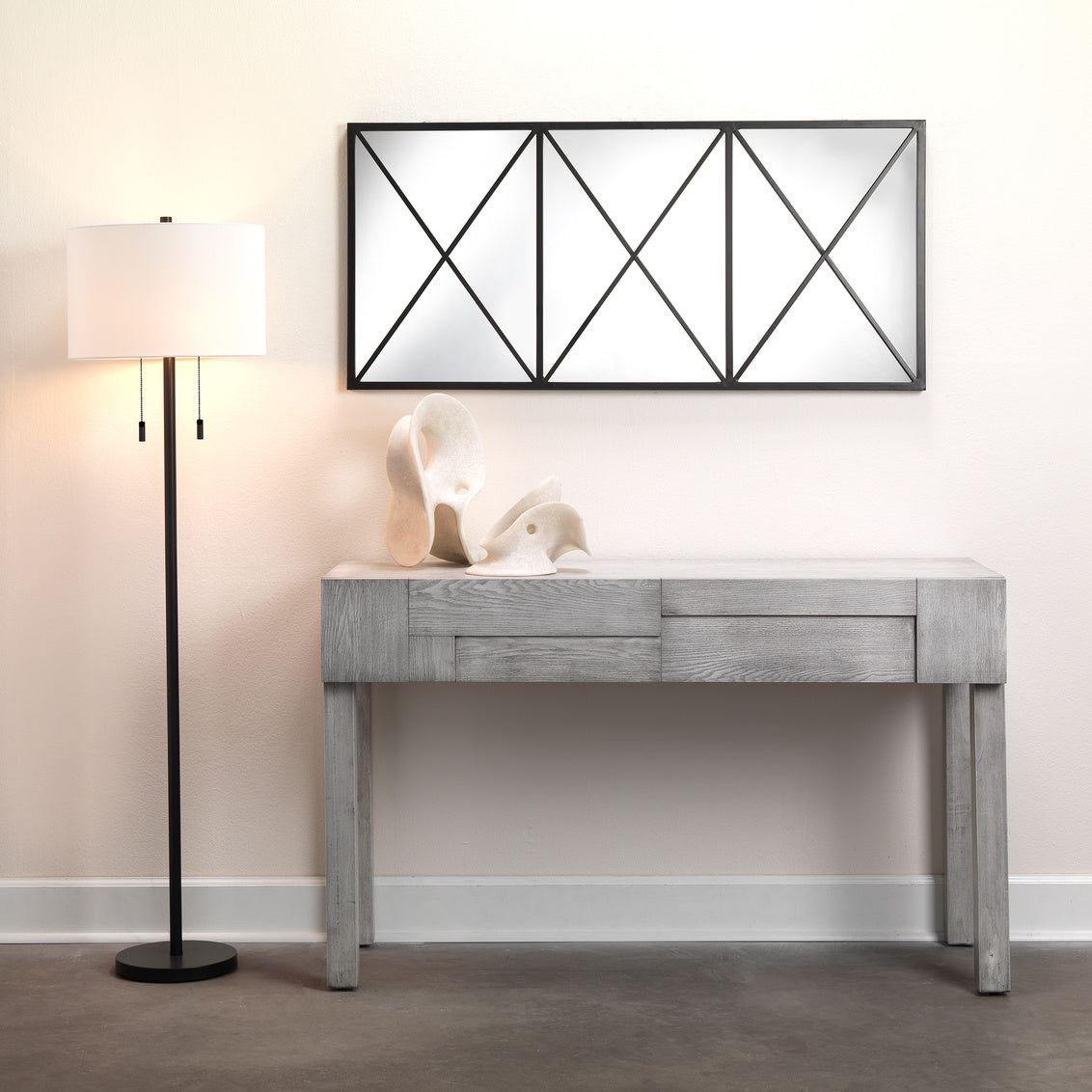 Gray console table with decorative items against a beige wall with a geometric mirror.
