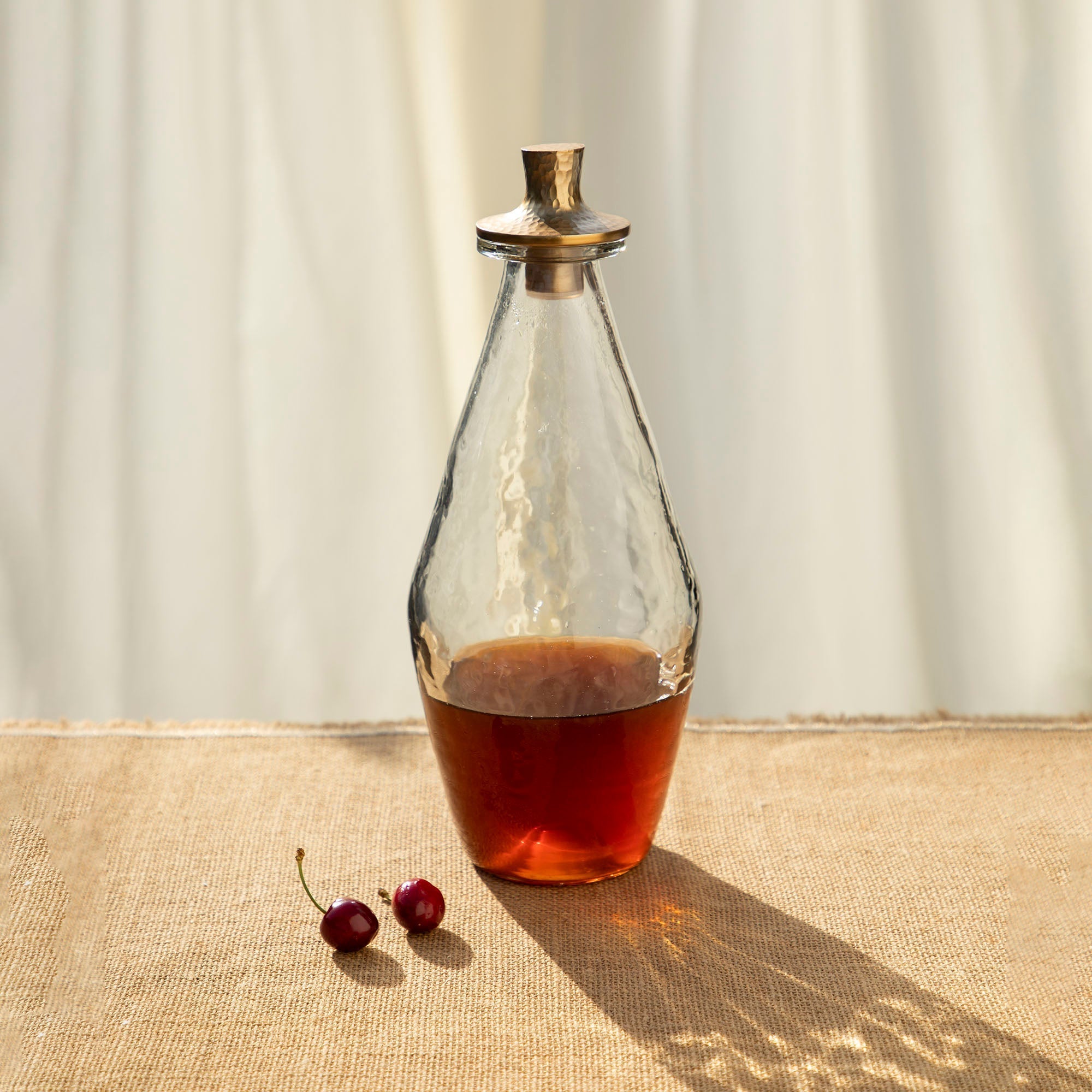 Pebbled glass decanter with a hammered brass cap on a textured surface with two cherries.