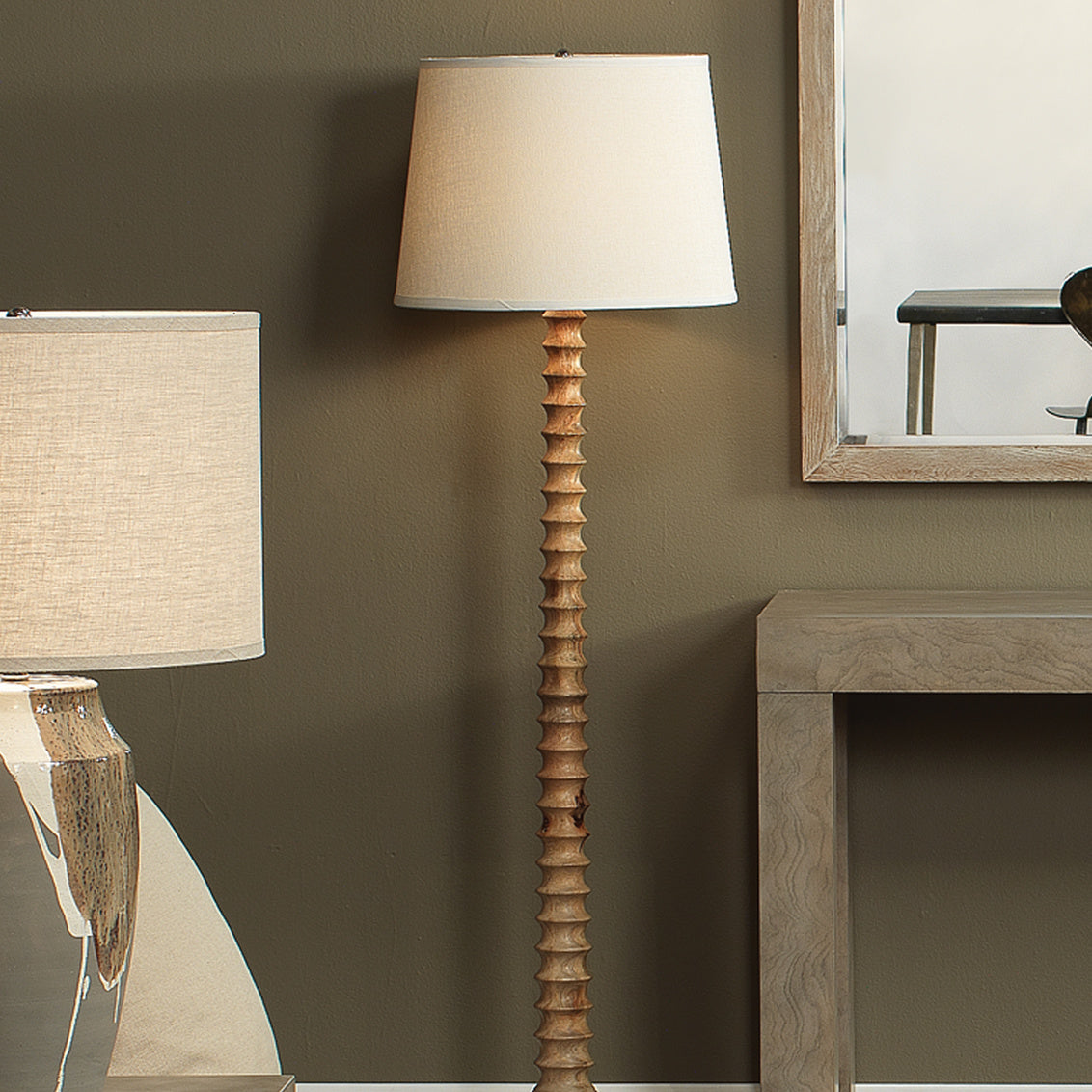 Wooden floor lamp with a white shade next to a mirror on a wall.