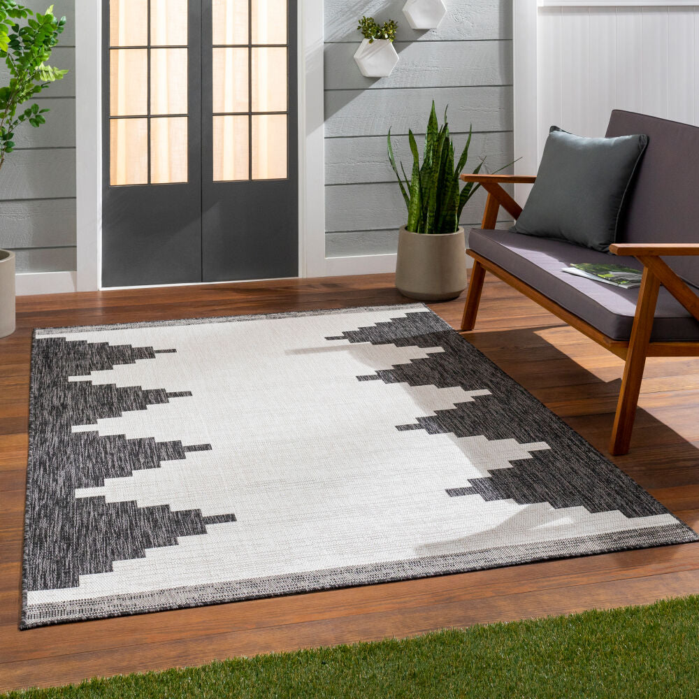 Geometric patterned rug on a wooden floor with a chair and plants in the background.