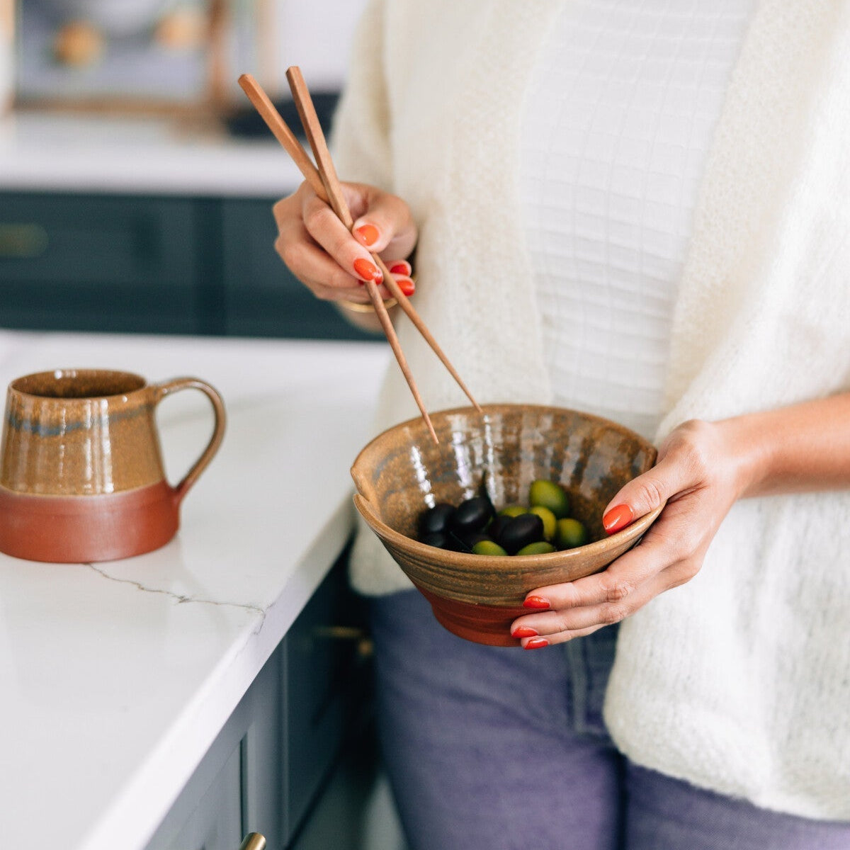 Person holding a bowl of olives with chopsticks in a kitchen setting