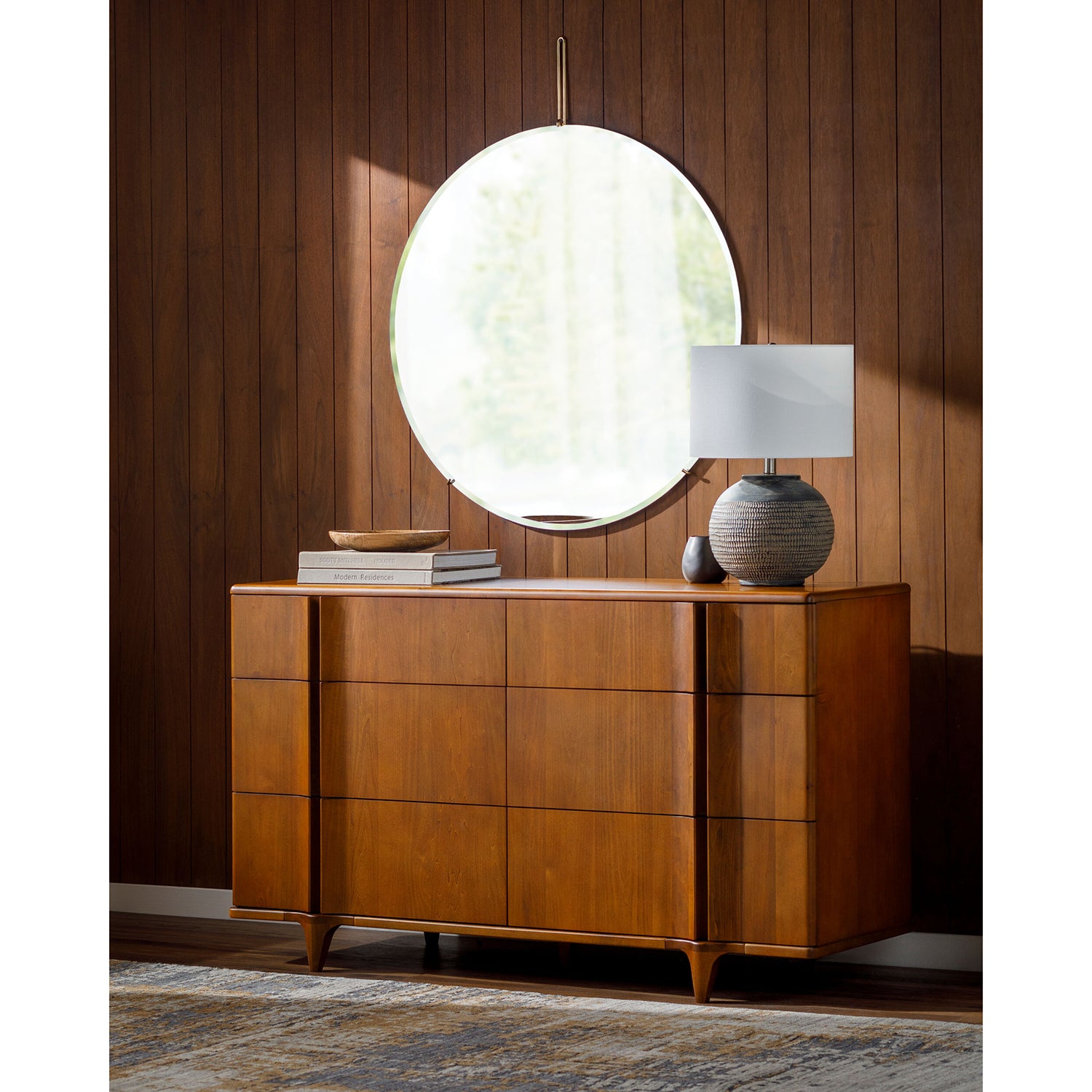 Wooden dresser with round mirror and lamp against a wooden wall.
