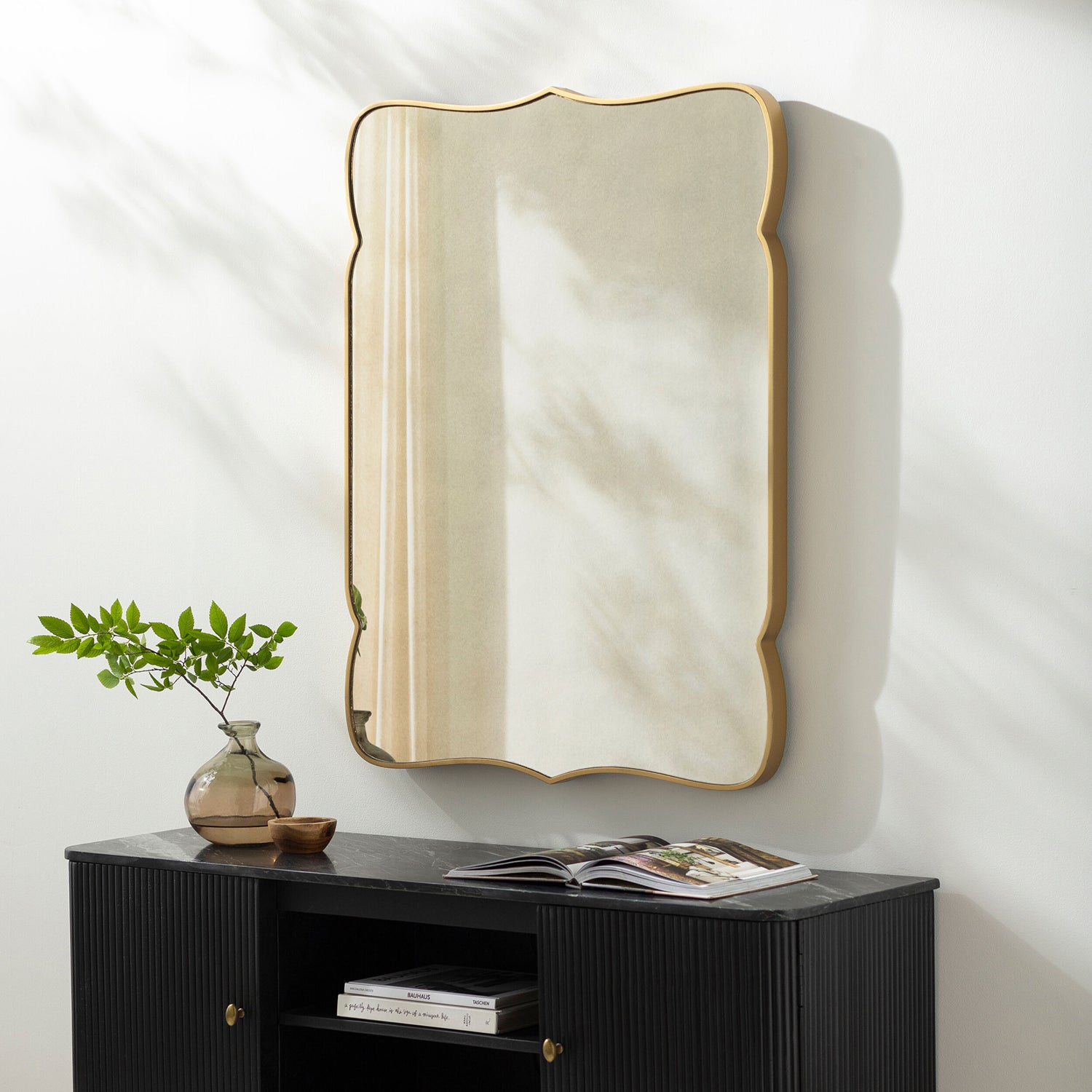 Decorative mirror with gold frame on a white wall above a black cabinet with books and a plant.