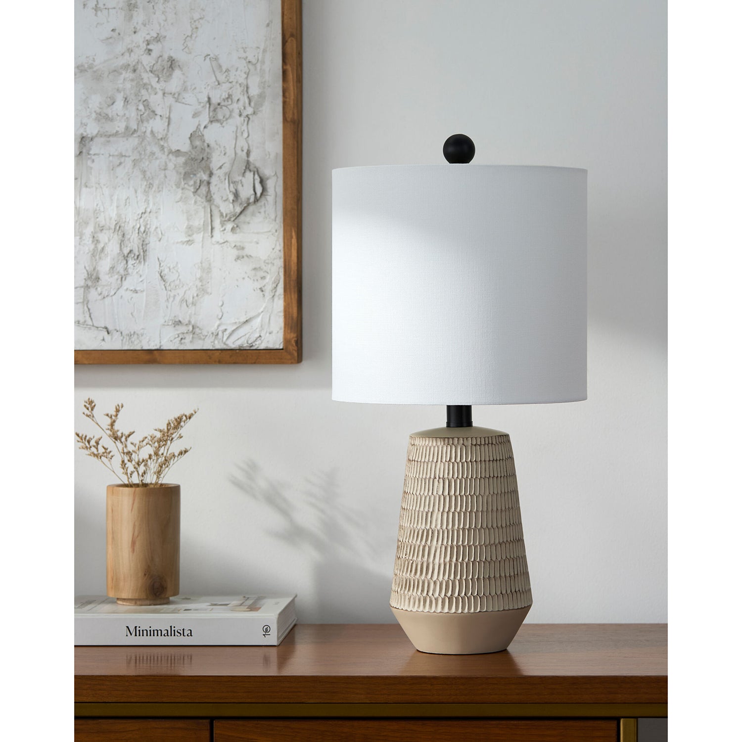 Beige textured table lamp on a wooden surface with a white wall and framed abstract art in the background.