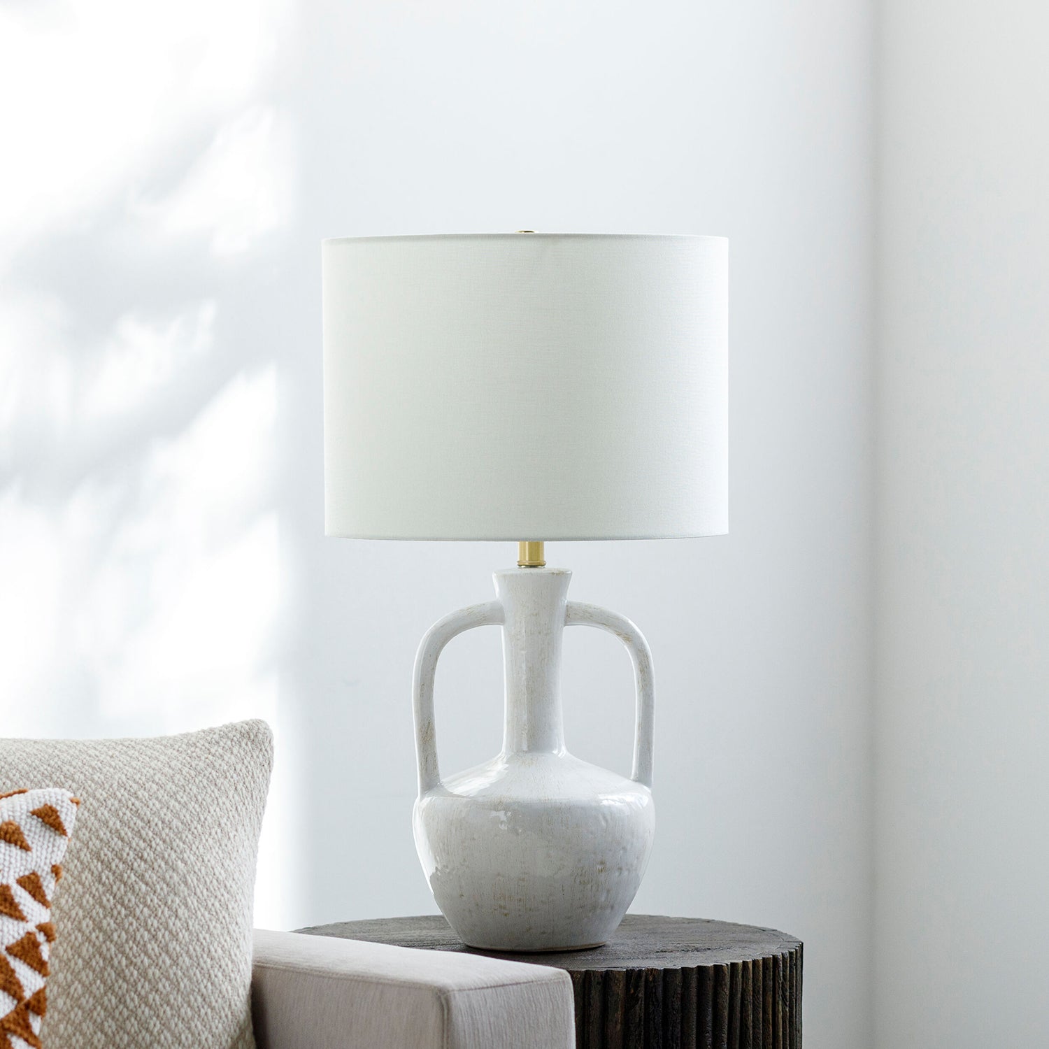 White ceramic lamp with a beige lampshade on a wooden side table against a white wall.