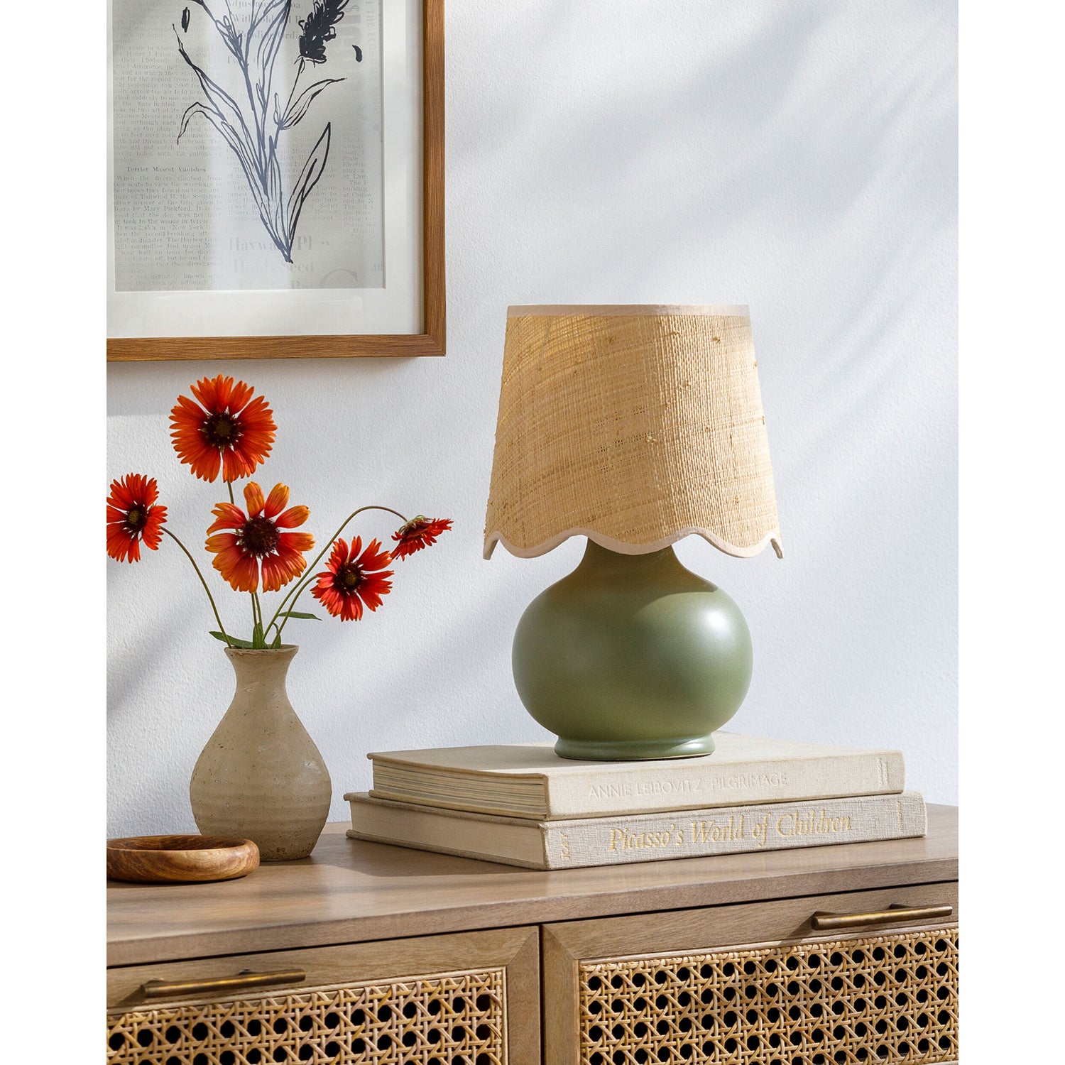 Green lamp with woven shade on a wooden sideboard with books and flowers.
