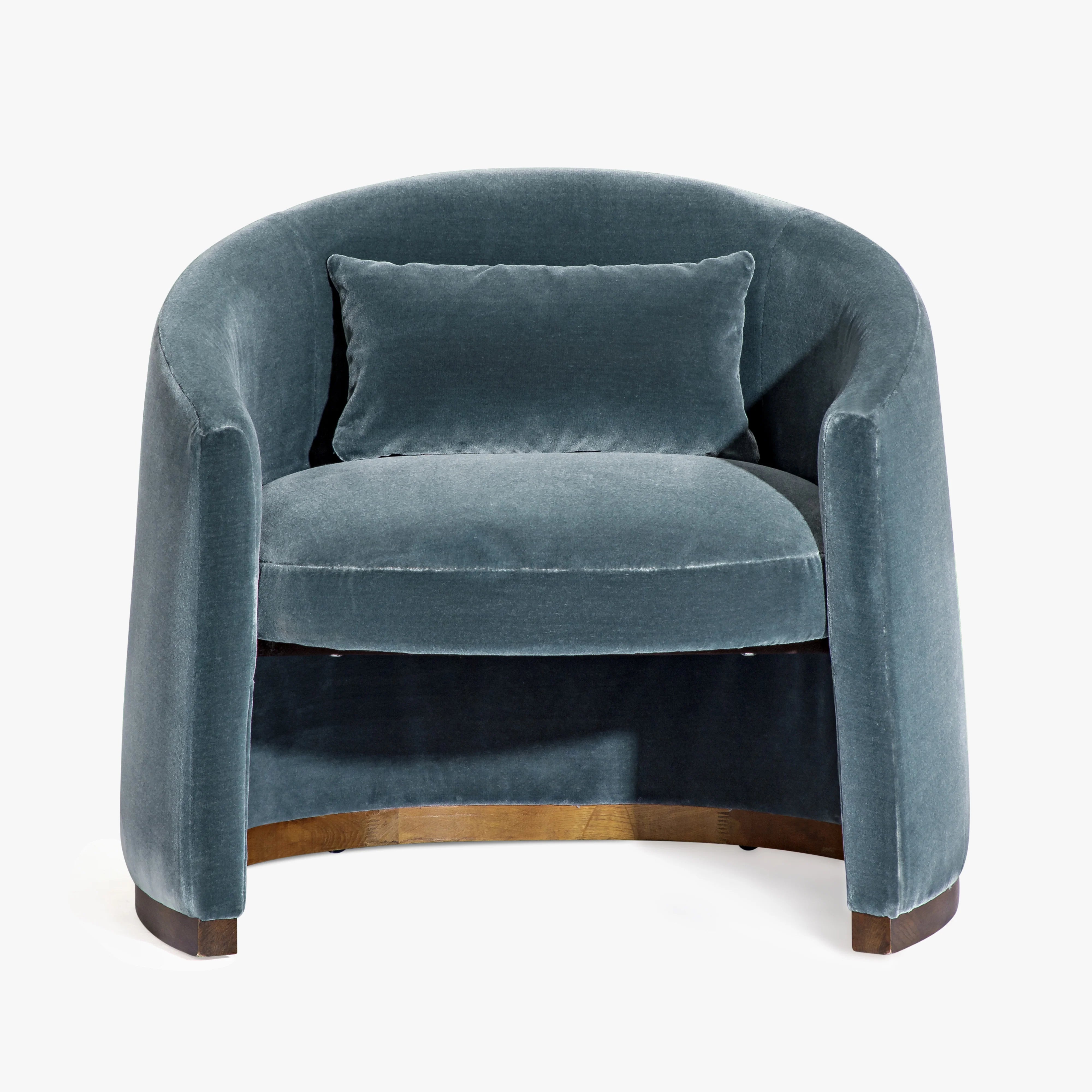 A modern barrel-style armchair upholstered in textured blue-gray velvet with a matching throw pillow, featuring a warm wood base and dark wooden feet, set against a plain white background.