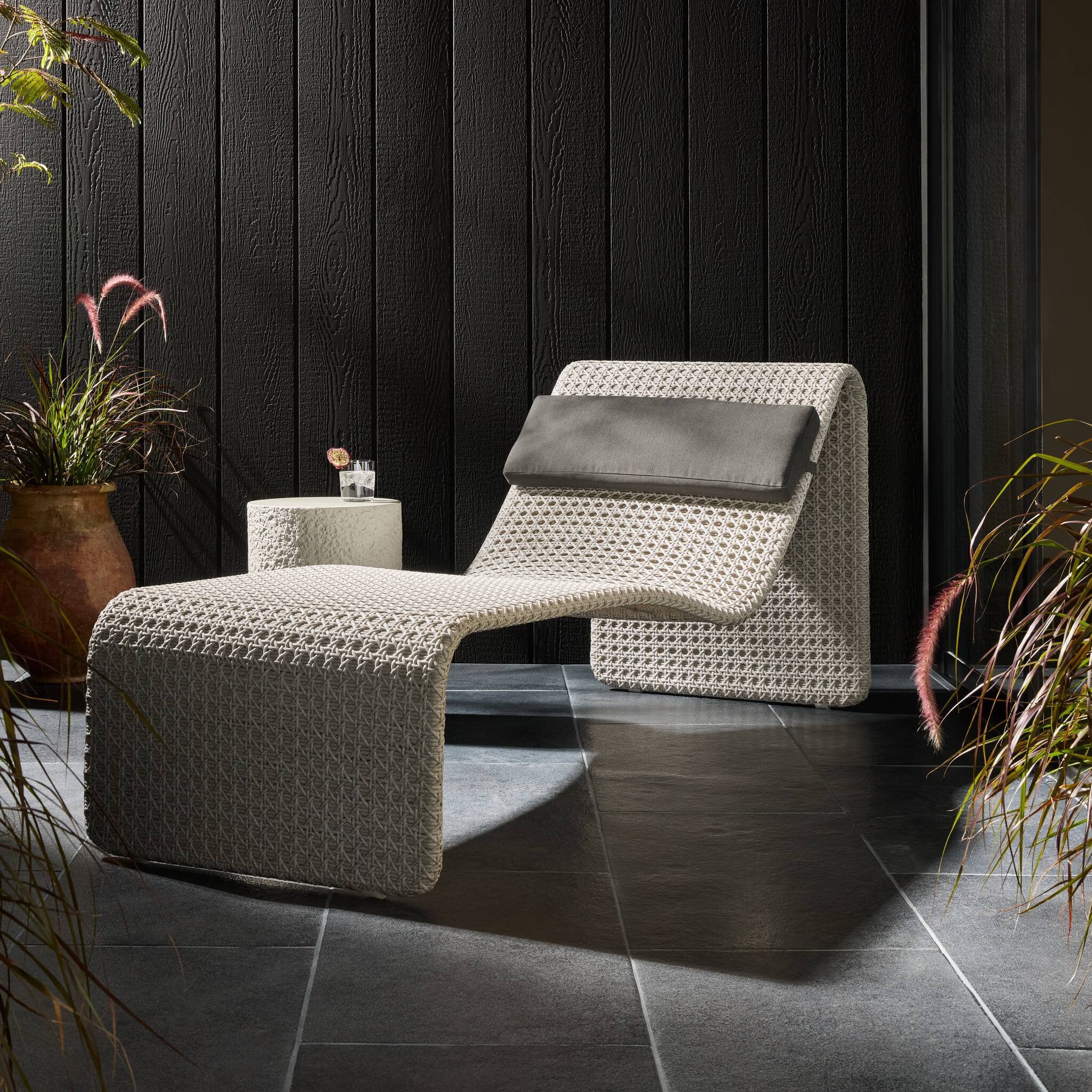 Modern outdoor lounge chair with ottoman on a dark tiled floor with plants.