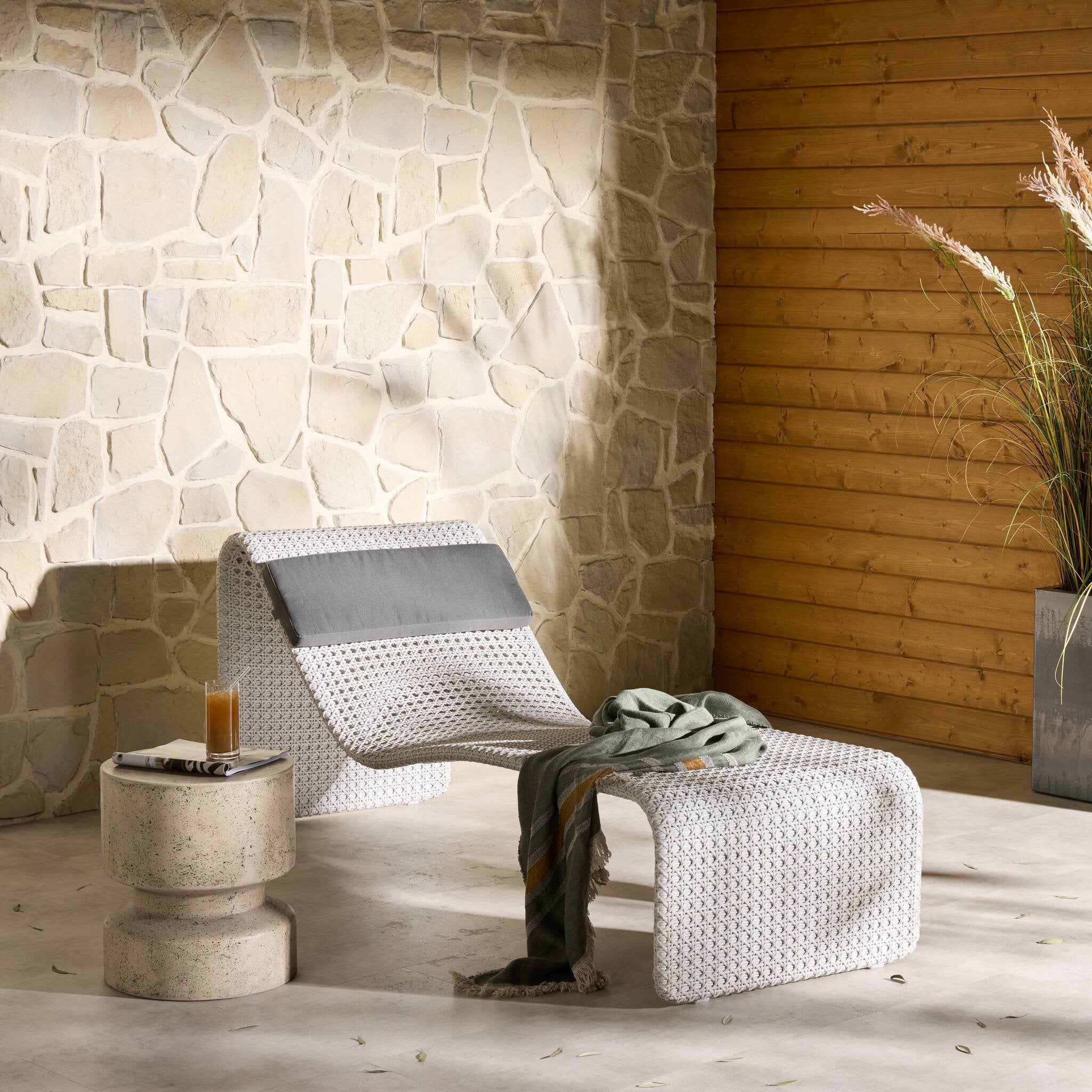 Cozy corner with a wicker chair, ottoman, and small table against a stone wall.