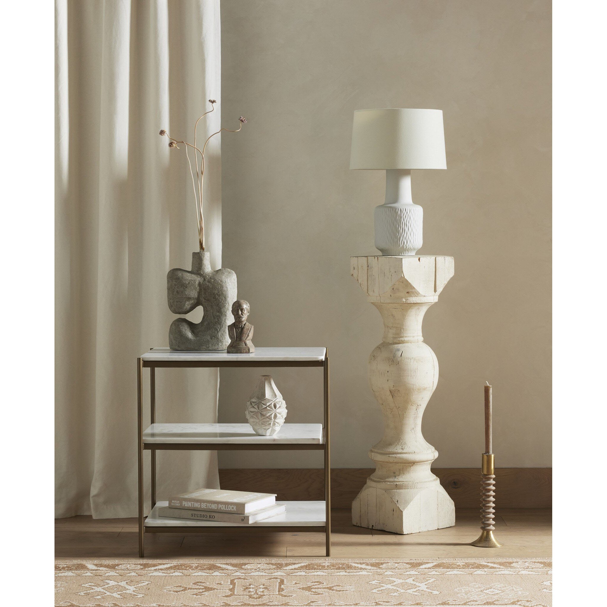 A styled interior scene with a tall white pedestal table holding a white lamp, next to a small metal-framed shelf with books and decor, set against a warm beige wall and cream curtains.