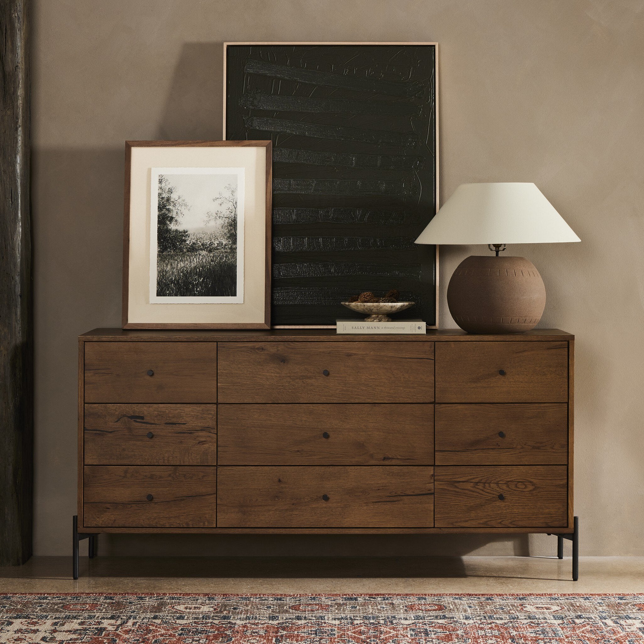 Wooden dresser with framed picture, lamp, and decorative items against a beige wall.