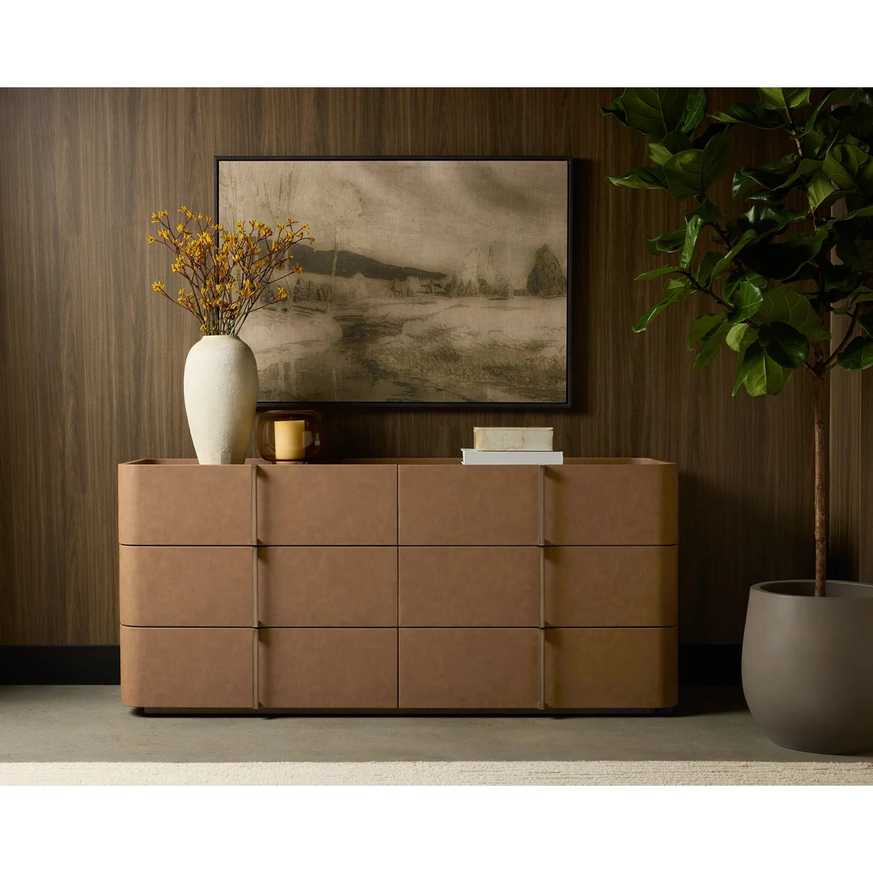 A modern 6-drawer dresser in warm tan leather, styled in a room setting with a white vase of yellow flowers, a framed landscape print, and a potted plant against a dark wood-paneled wall.