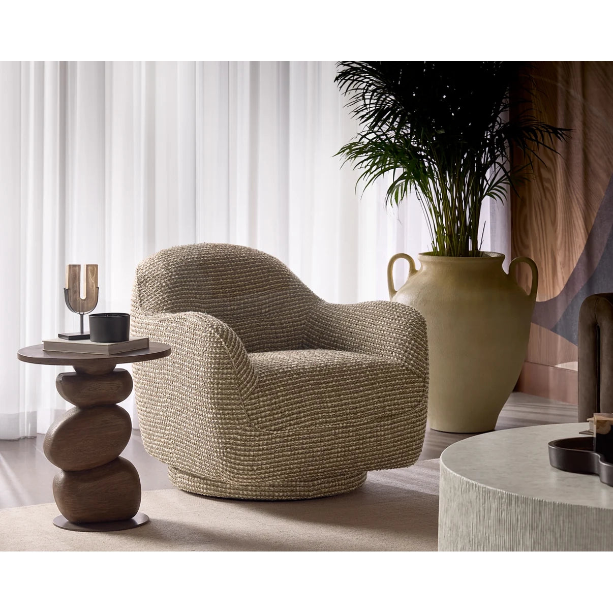 A modern round swivel armchair upholstered in a textured beige woven fabric, styled in a minimalist living room setting next to a stacked wooden side table and a large potted plant.
