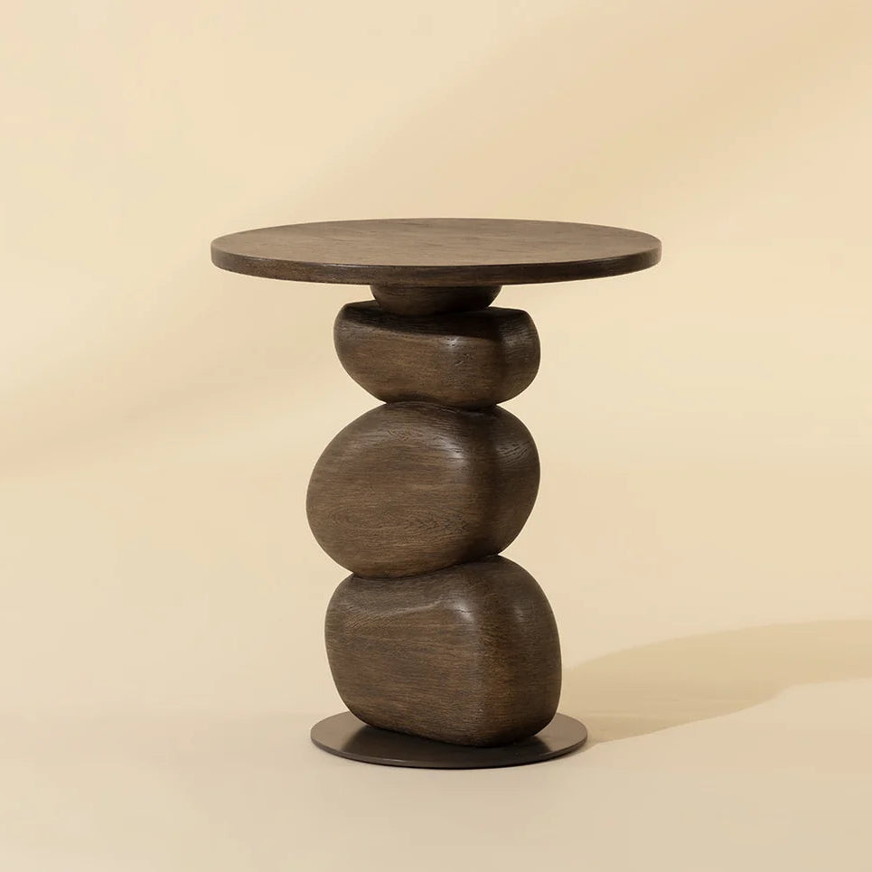 A dark brown wood-like side table with a round top and a stacked, organic-shaped pedestal base, set against a warm beige background.