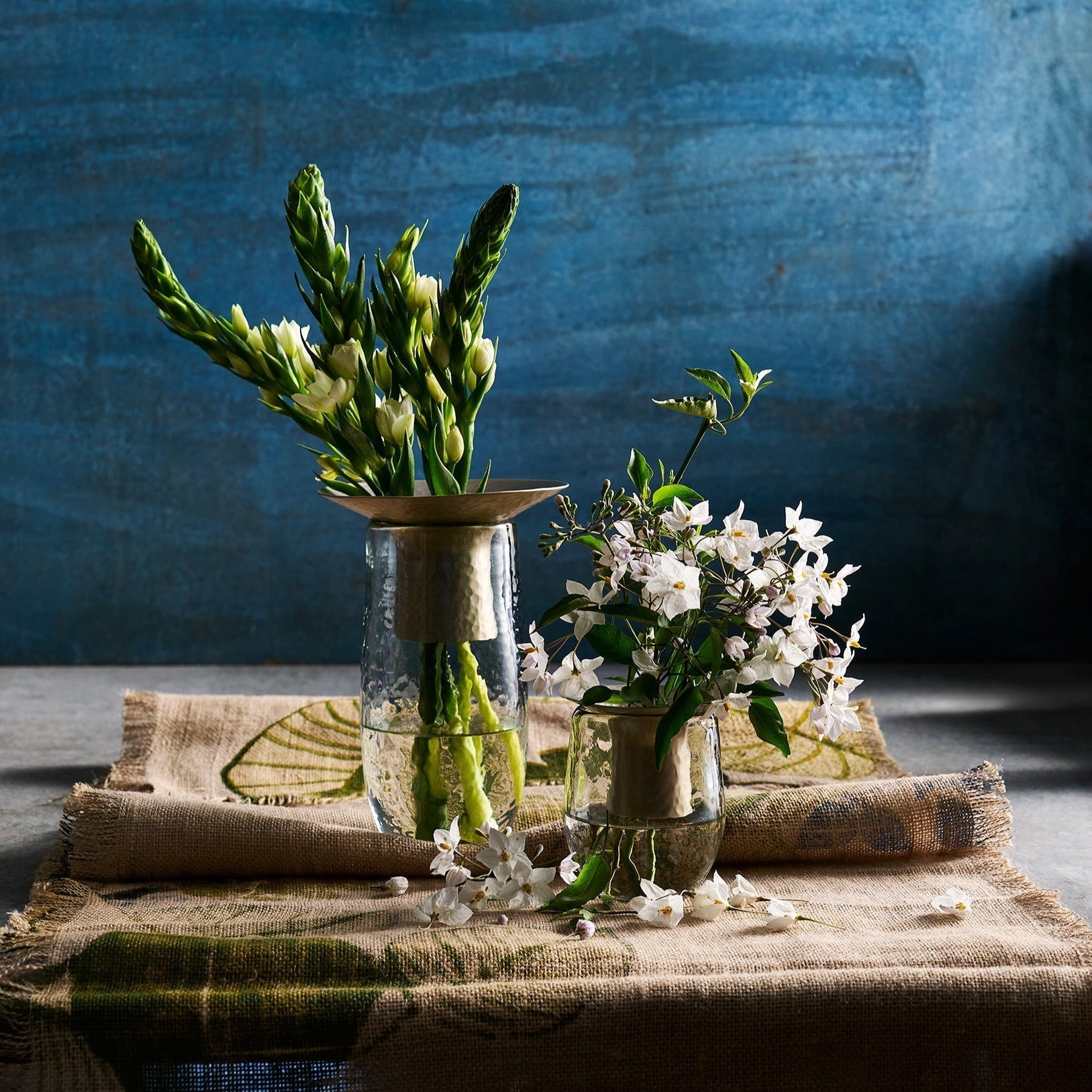 Two glass vases with greenery and white flowers on a textured tablecloth against a blue wall.