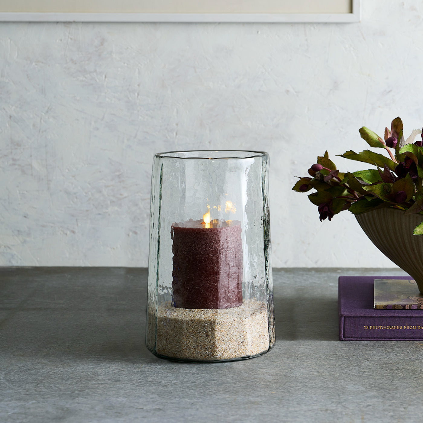 Glass candle holder with a red candle on a gray surface, with a plant and books in the background.
