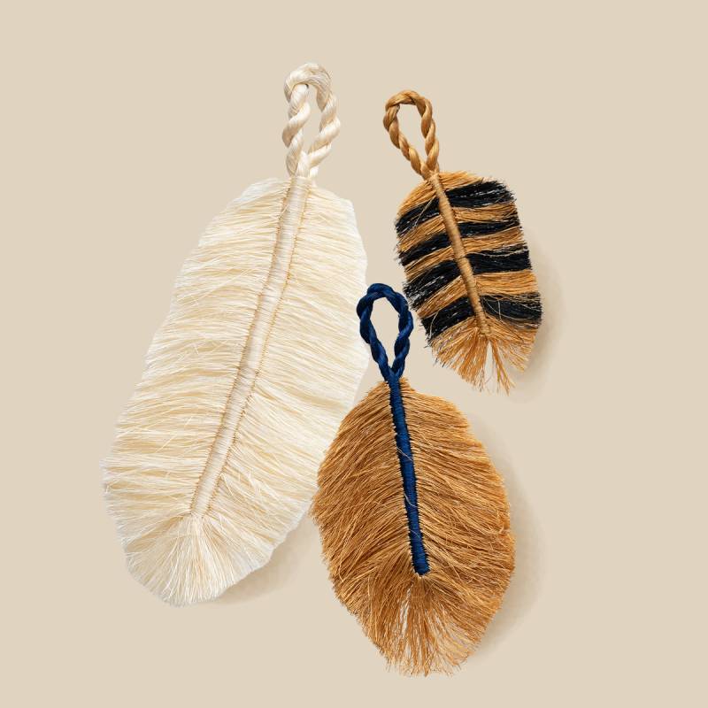 Three feather-shaped decorative bag charms with rope handles on a beige background