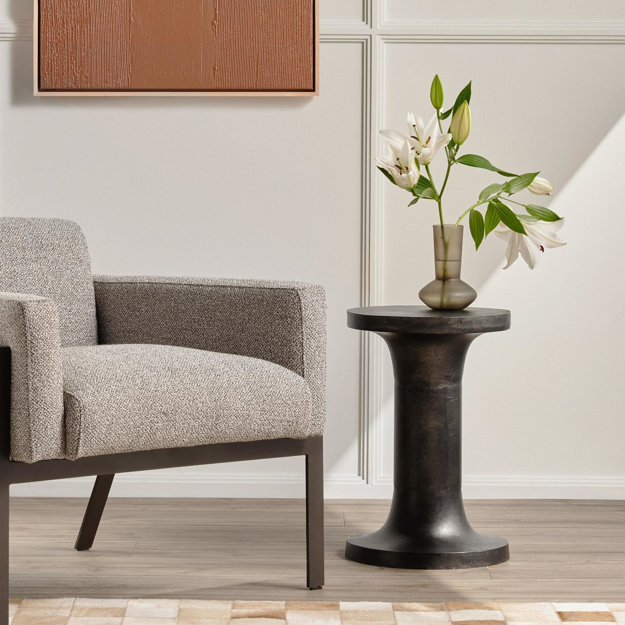 Gray armchair next to a black side table with a vase of flowers in a room with wooden flooring and a neutral wall.