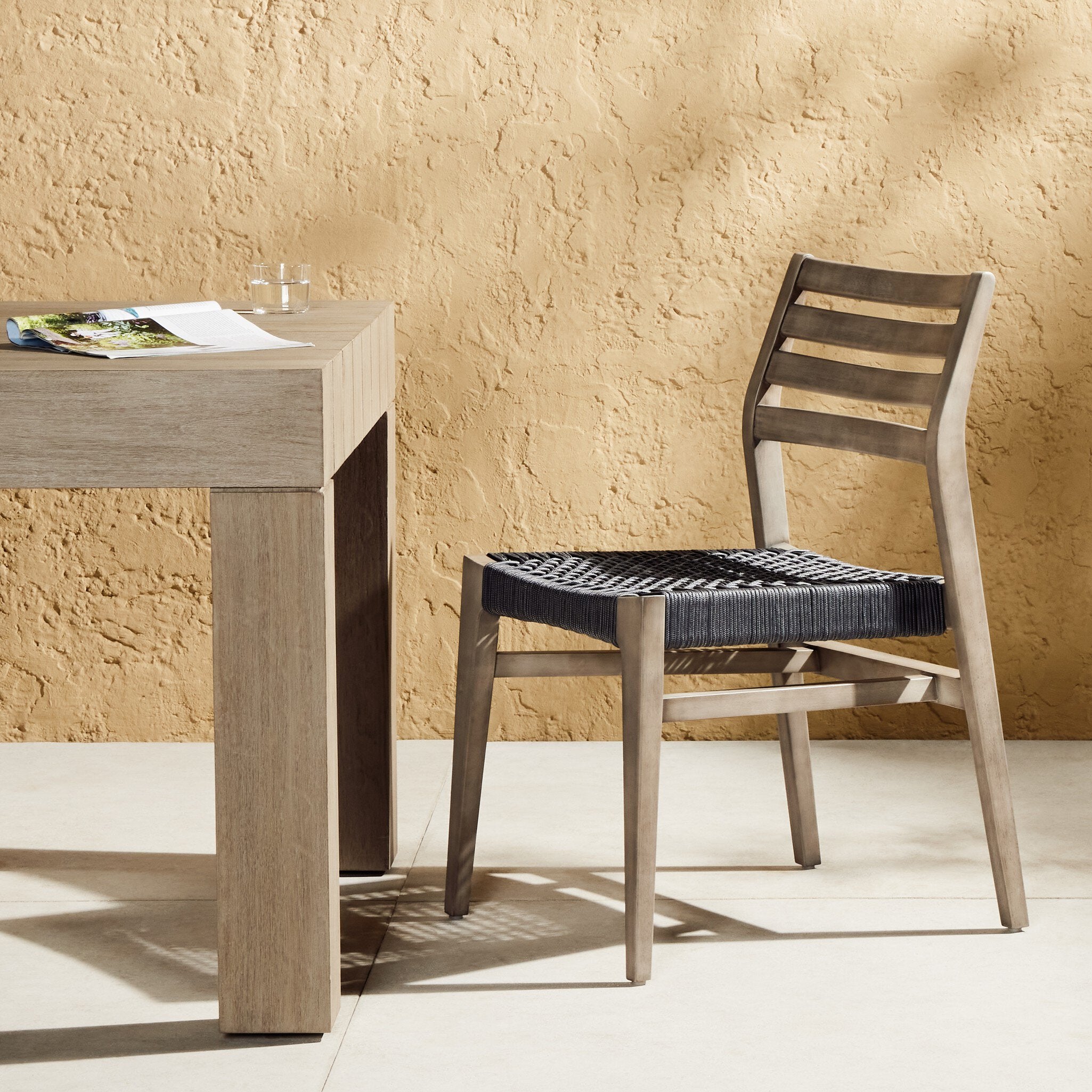 Wooden outdoor dining chair with a woven seat next to a wooden table against a textured beige wall.