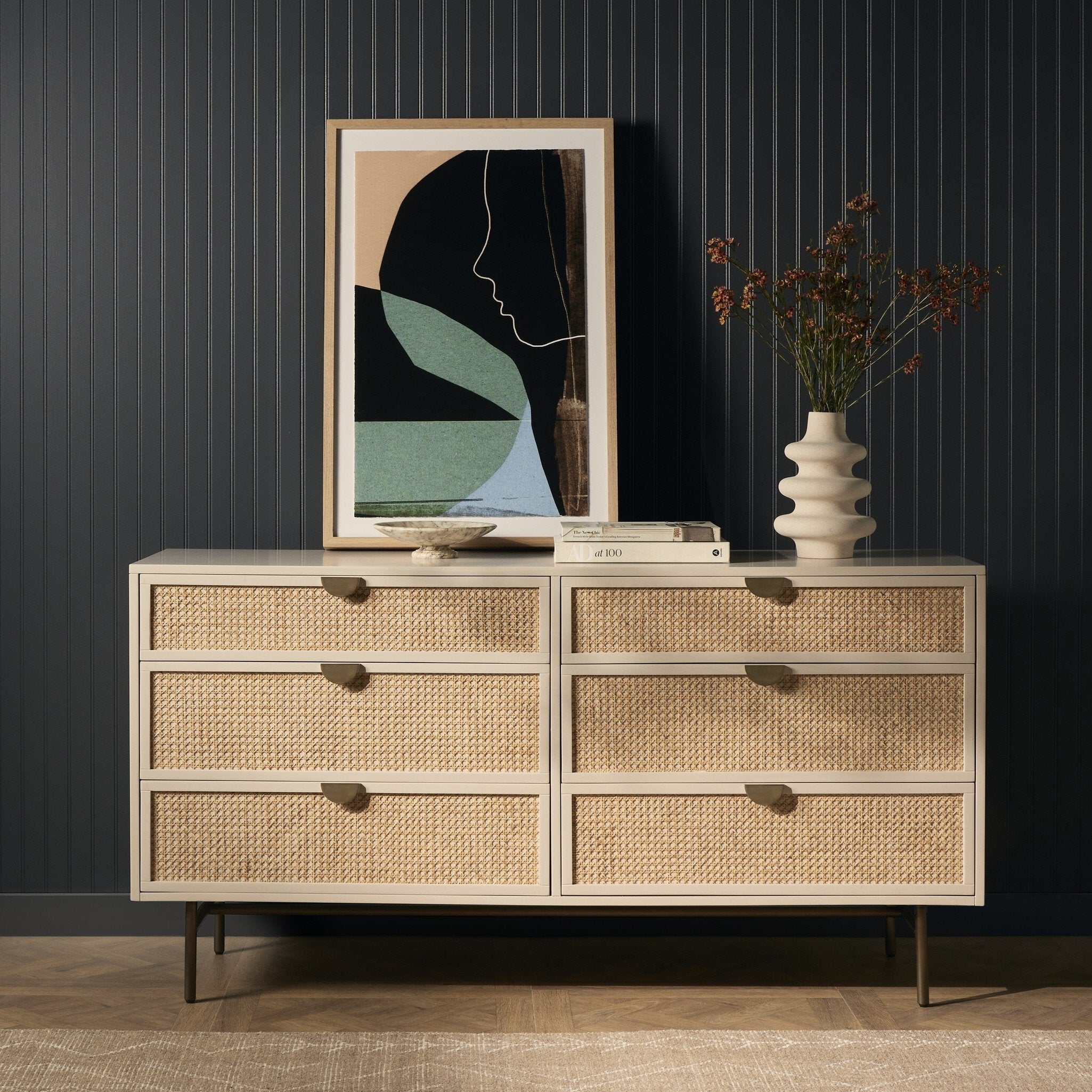 Beige dresser with woven drawers against a dark paneled wall, featuring a framed abstract art piece and a vase with flowers.