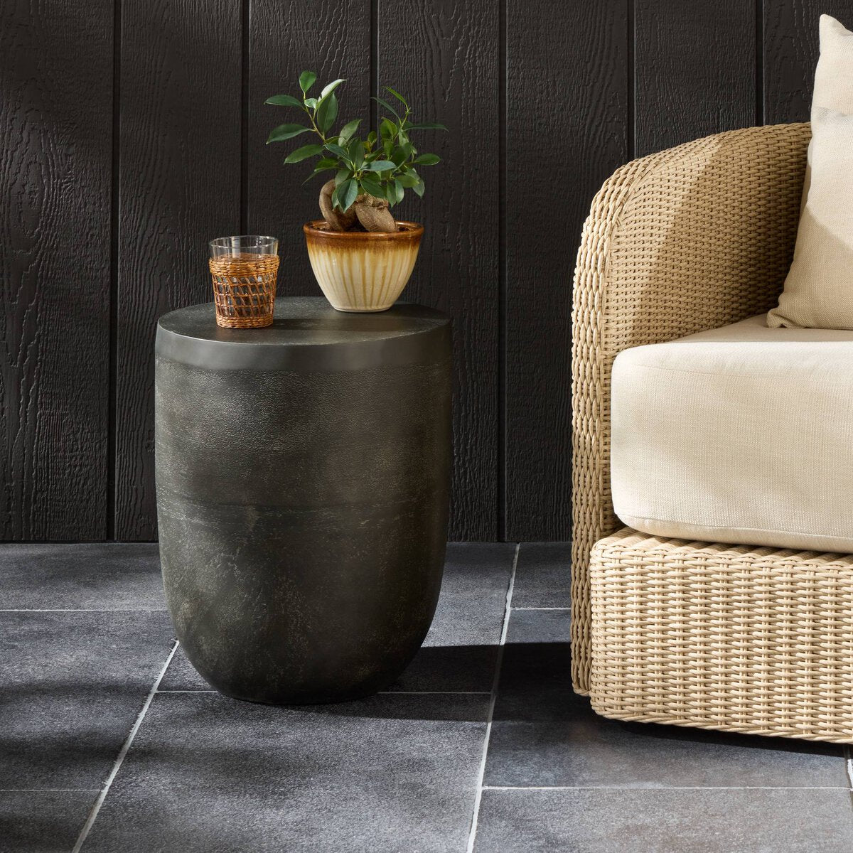 Aluminum side table with a plant and glass next to a wicker chair against a dark paneled wall.