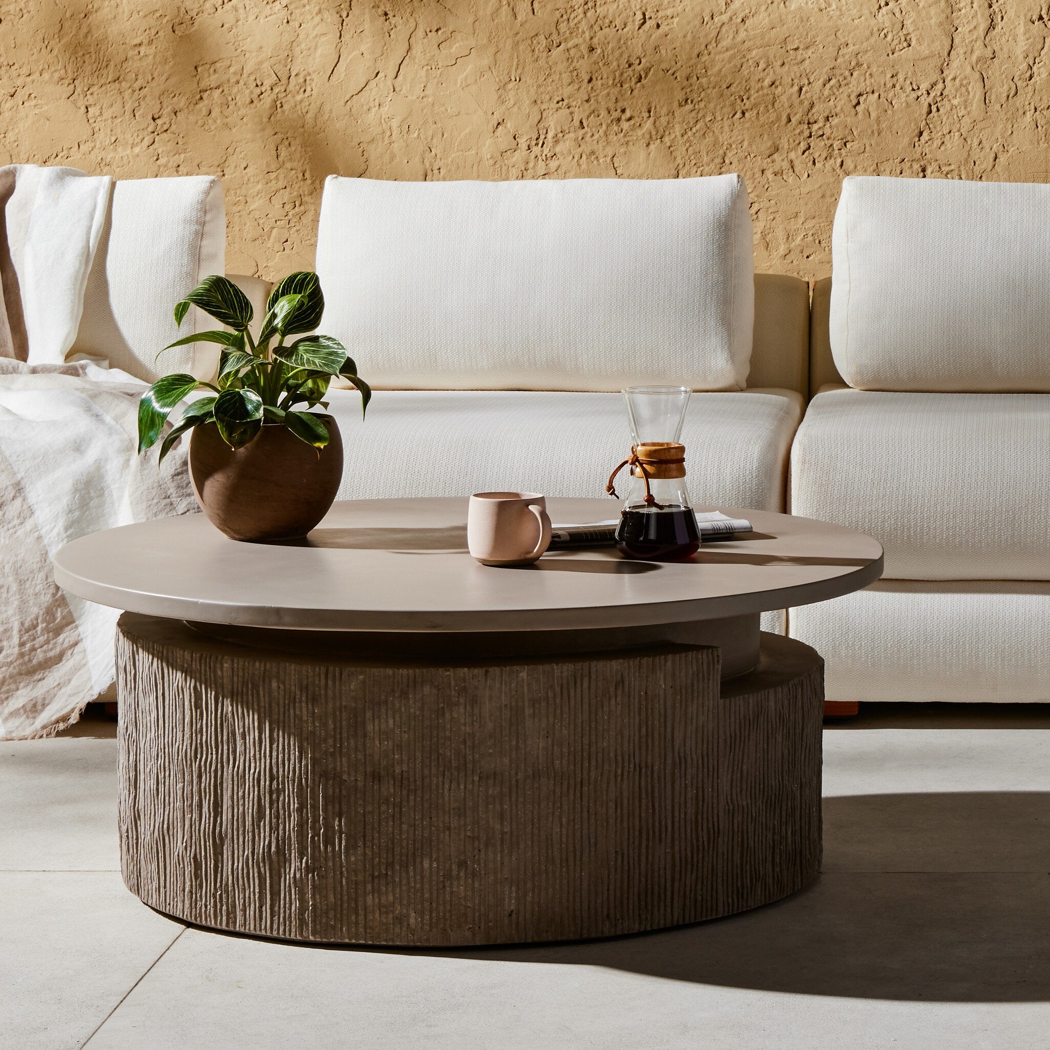 Round concrete coffee table with a plant, mug, and coffee maker in front of a white sofa against a beige wall.