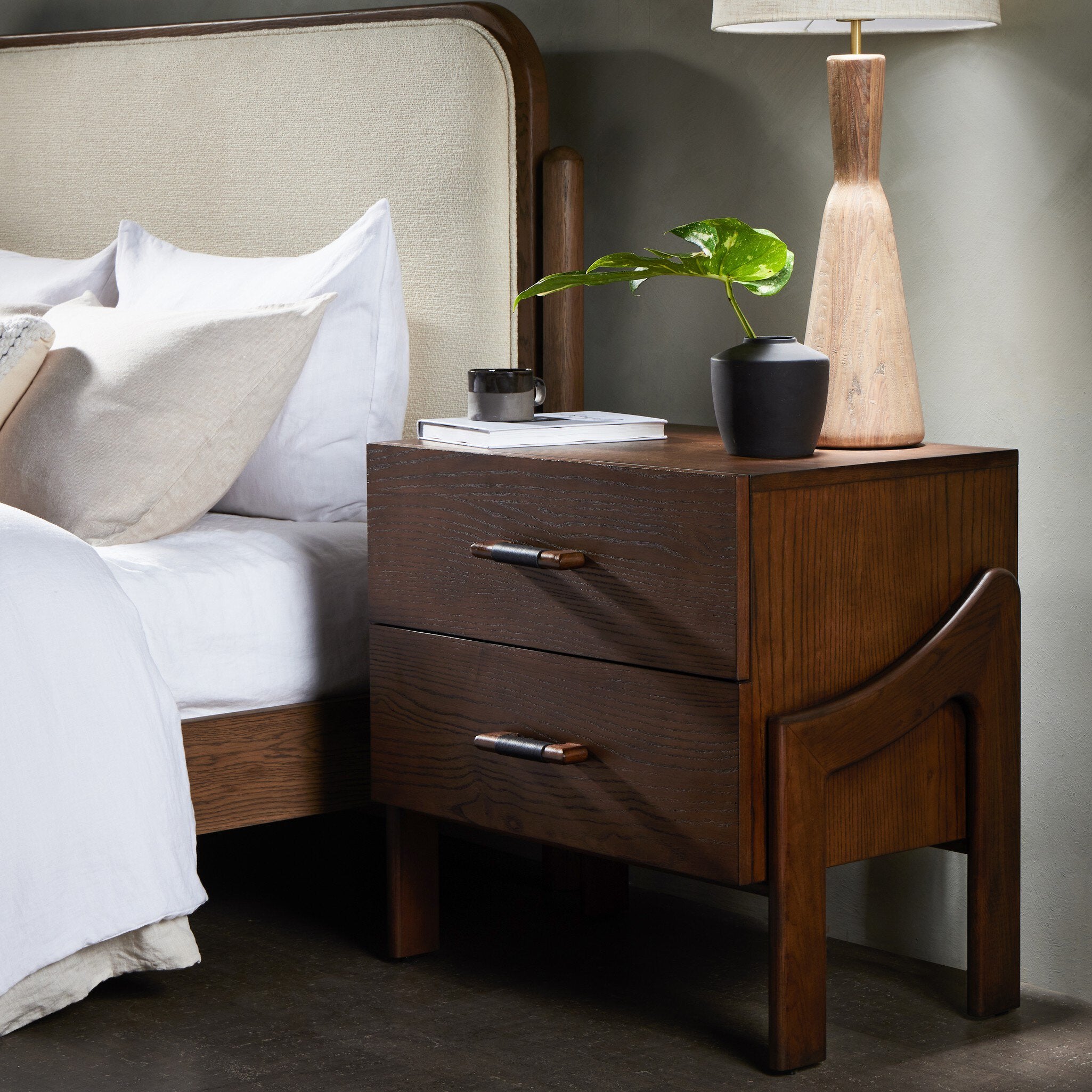 Wooden nightstand with lamp and plant next to a bed with white bedding.