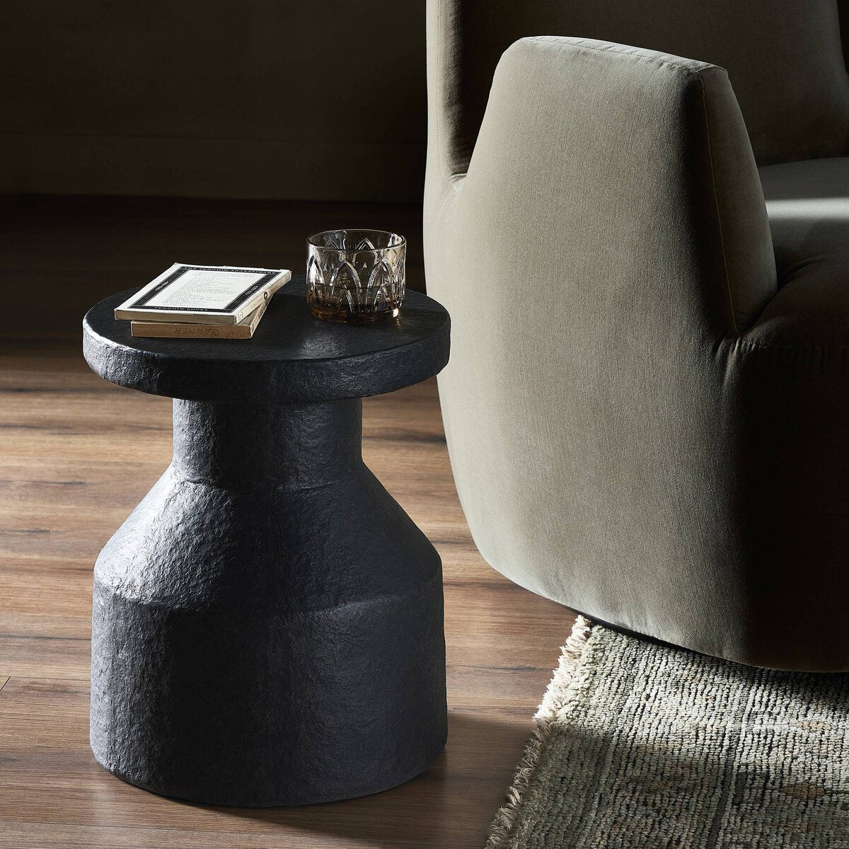 Black side table with a glass and book next to a gray armchair on a wooden floor.