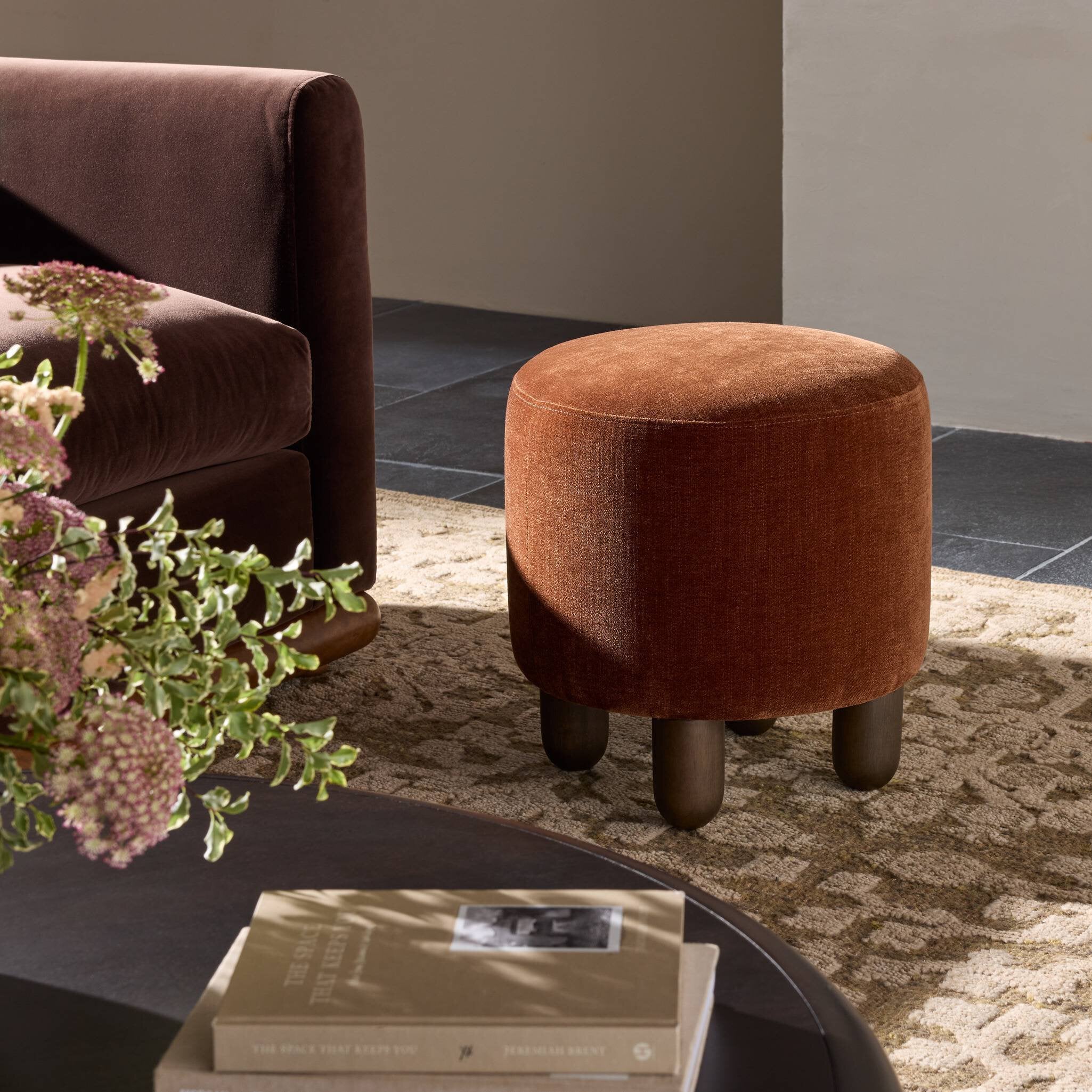 Brown ottoman next to a sofa with books and flowers on a coffee table in a living room setting.