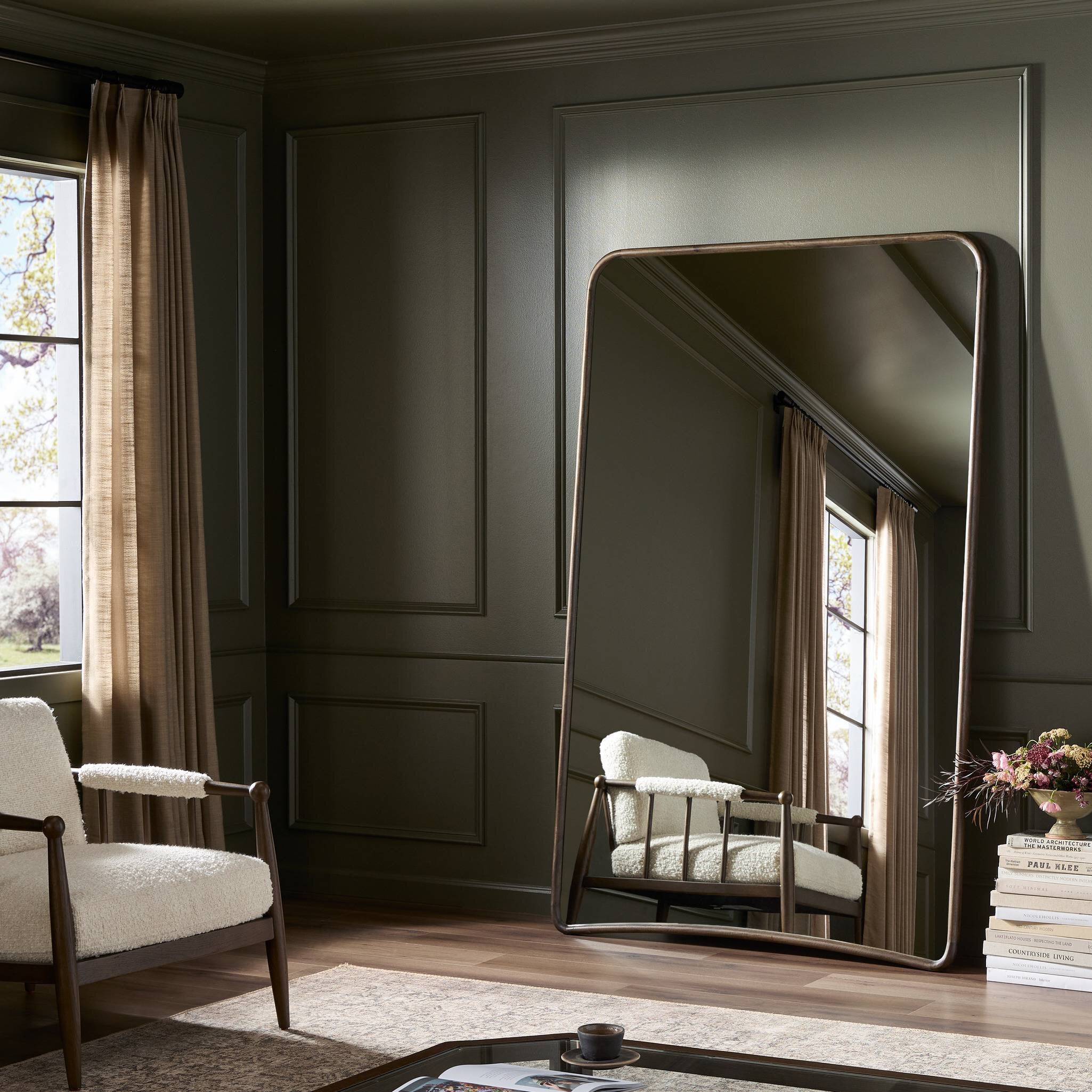 Large floor mirror reflecting a room with a chair and window.