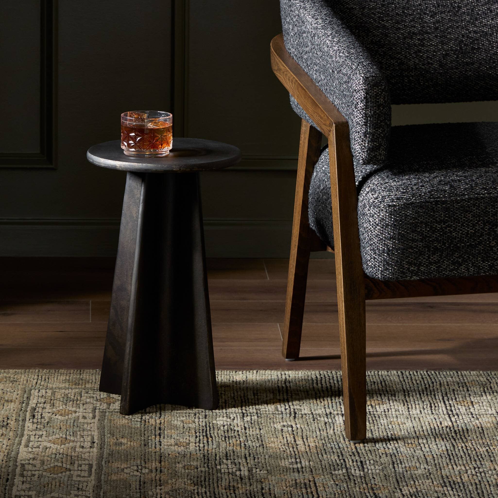 Small round black side table with two glasses of whiskey next to a gray upholstered chair.