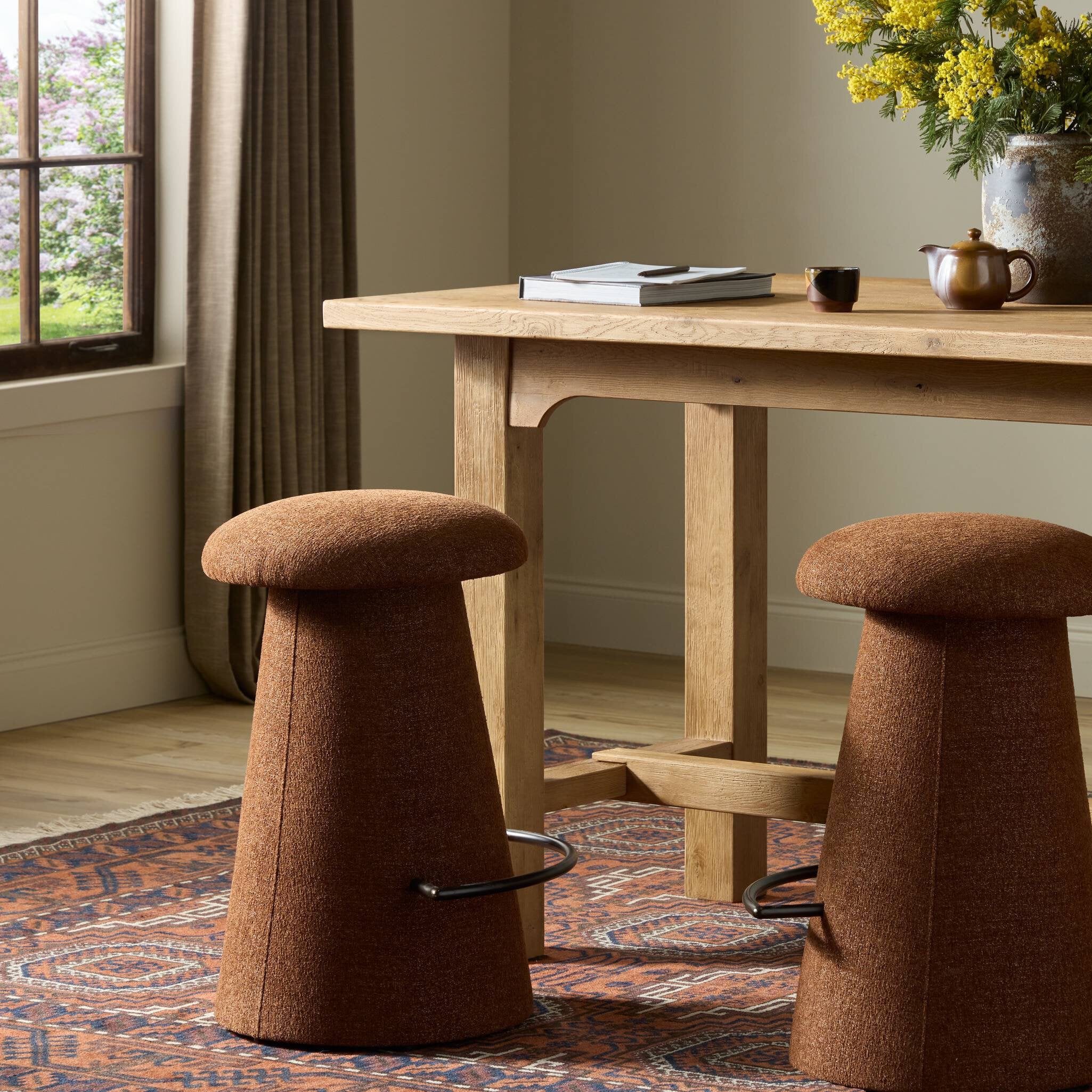 Two brown counter stools in front of a wooden table with a vase of flowers in a room with a window and rug.