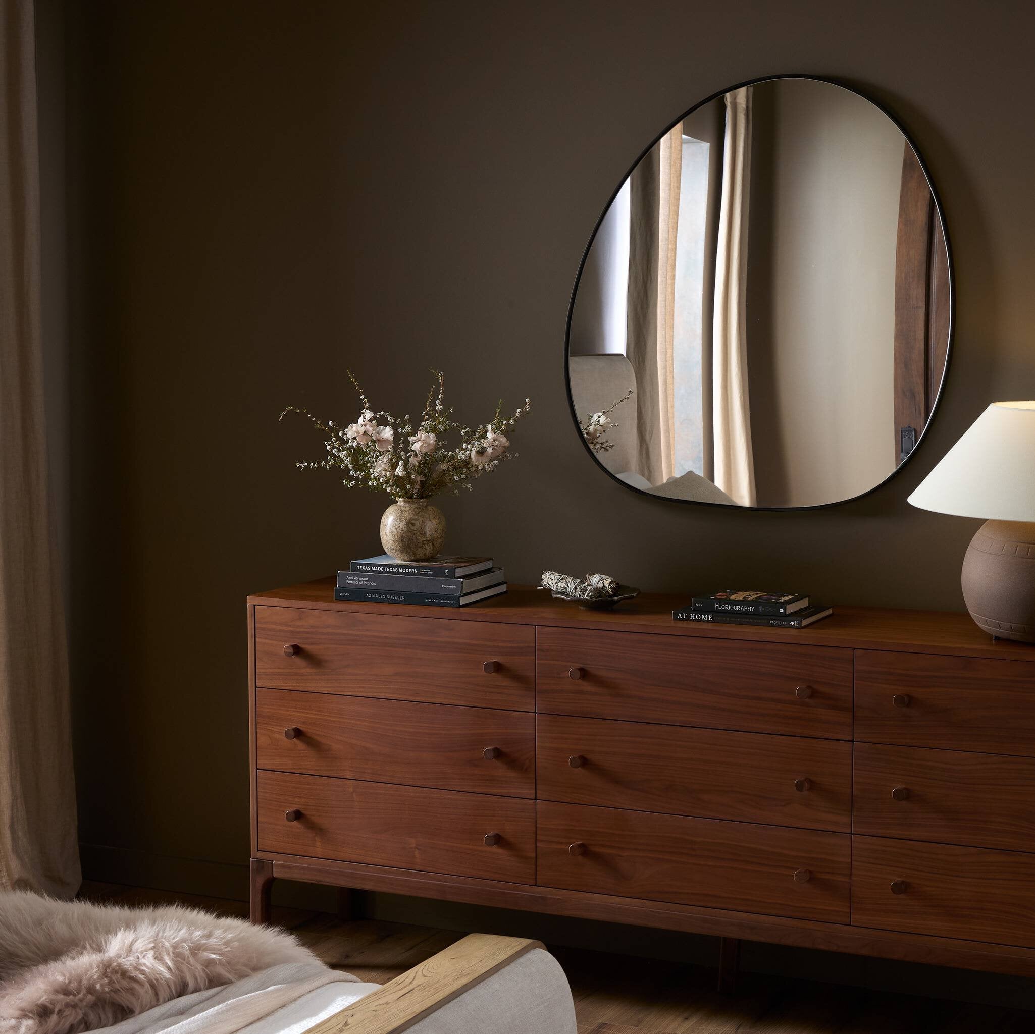 Wooden dresser with round mirror, lamp, and decorative items against a brown wall.