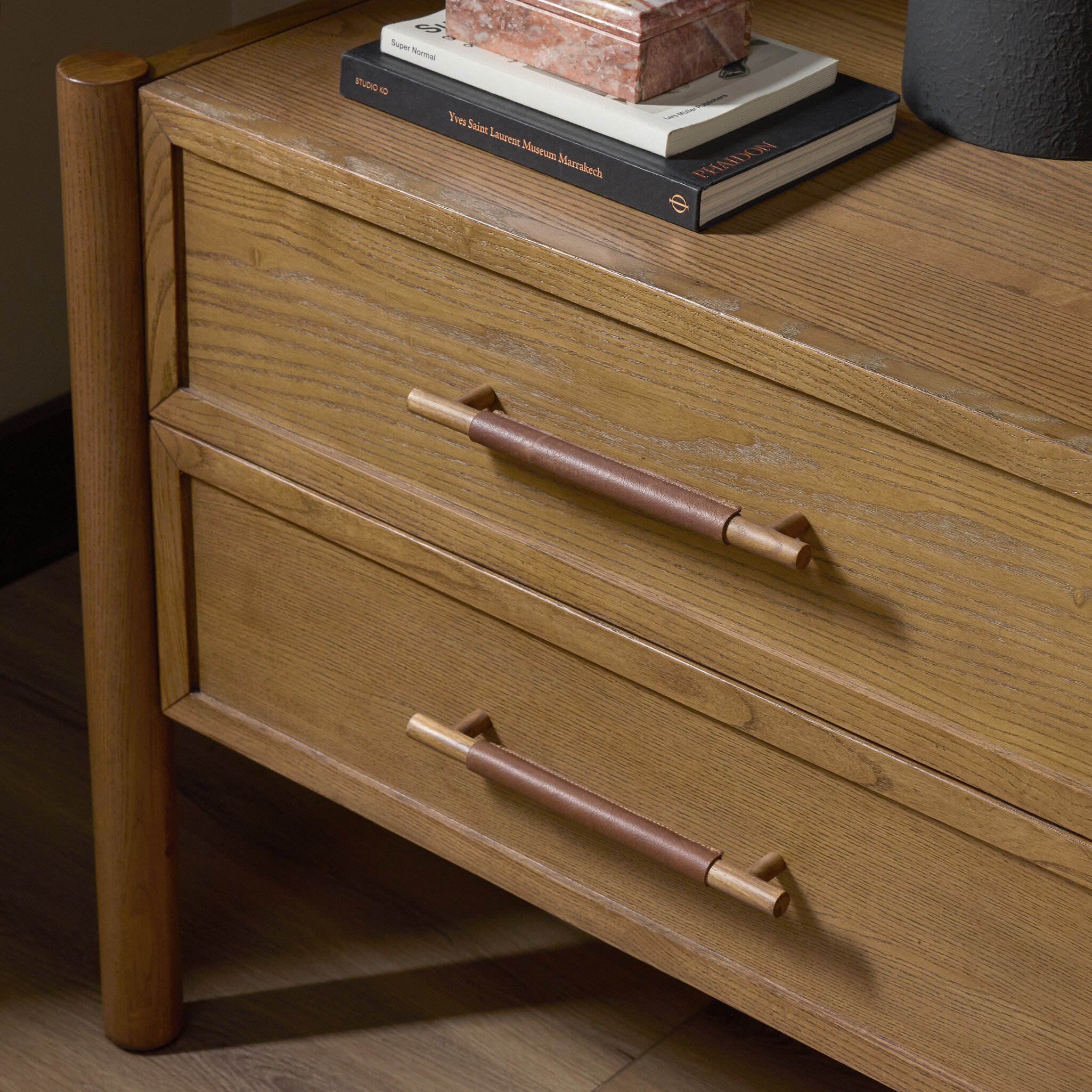 A close-up of a mid-century modern wooden side table or nightstand with two drawers, featuring warm medium-brown wood grain and cylindrical metal handles with a copper-toned center section. Books and a vase are styled on top.