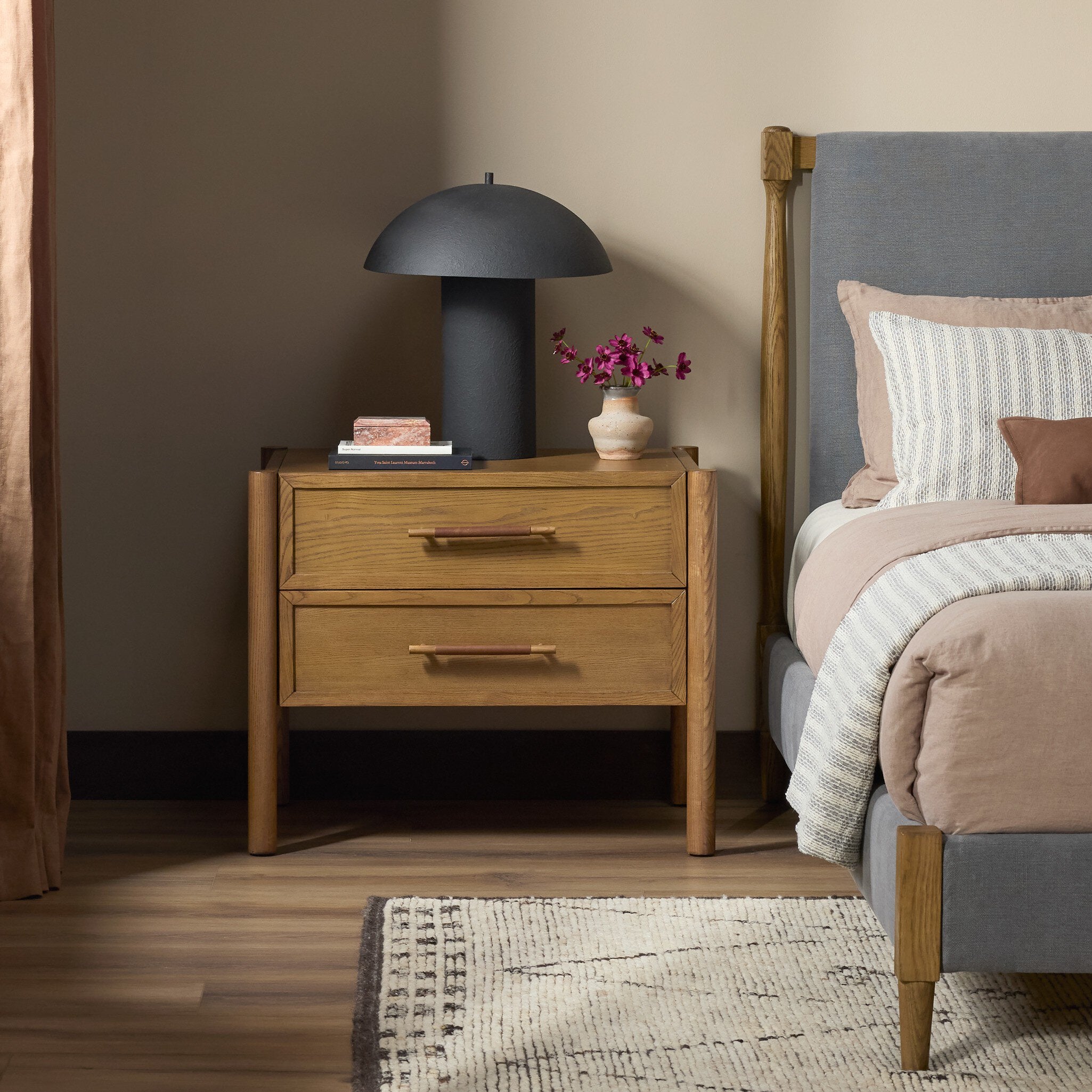 A two-drawer wooden nightstand in a warm medium oak finish, styled in a bedroom next to a bed with a gray upholstered headboard. On top of the nightstand is a matte black dome lamp and a small vase of pink flowers.