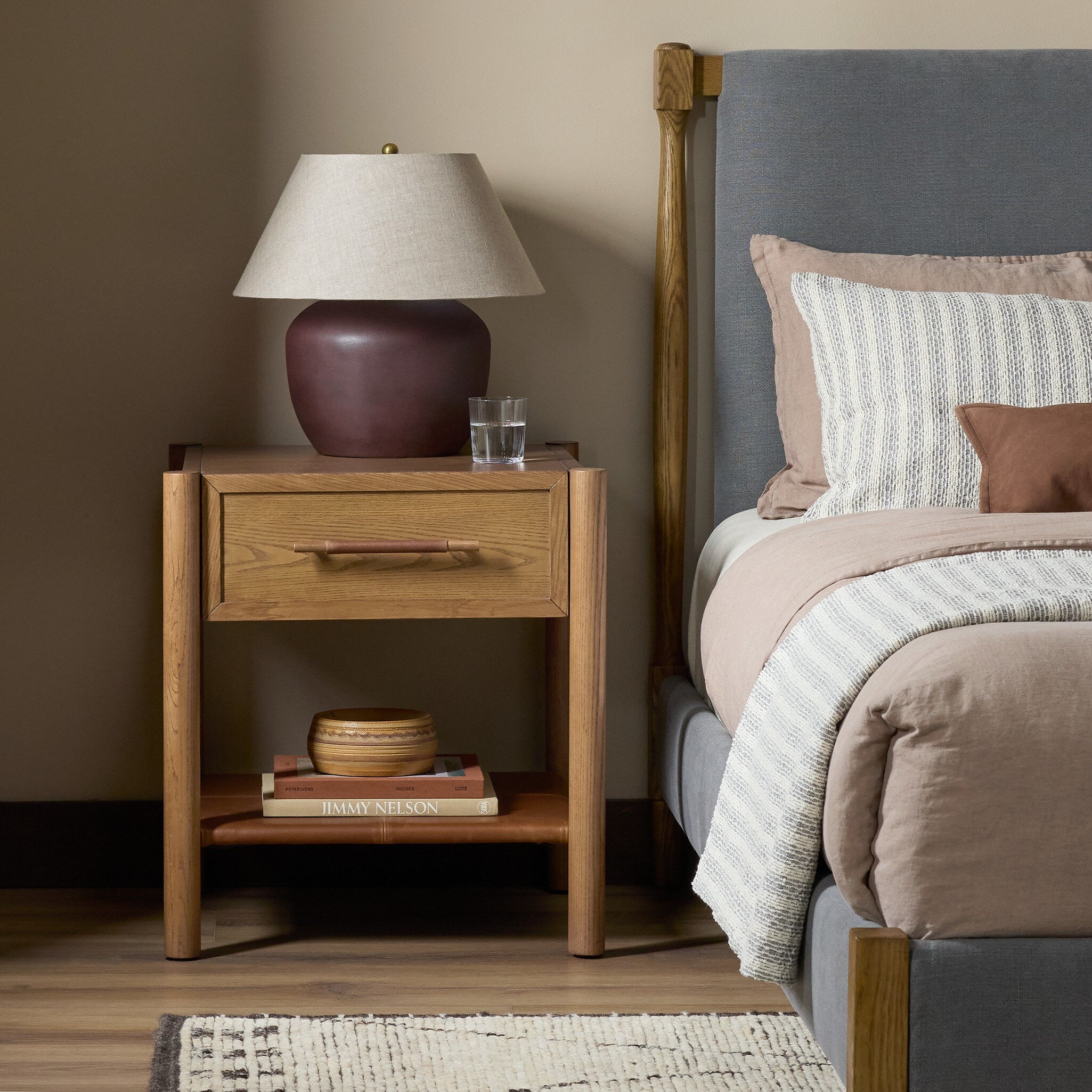 A mid-century modern wooden nightstand beside a bed with gray upholstered headboard and beige bedding, featuring a table lamp with a dark brown ceramic base and a beige drum shade.