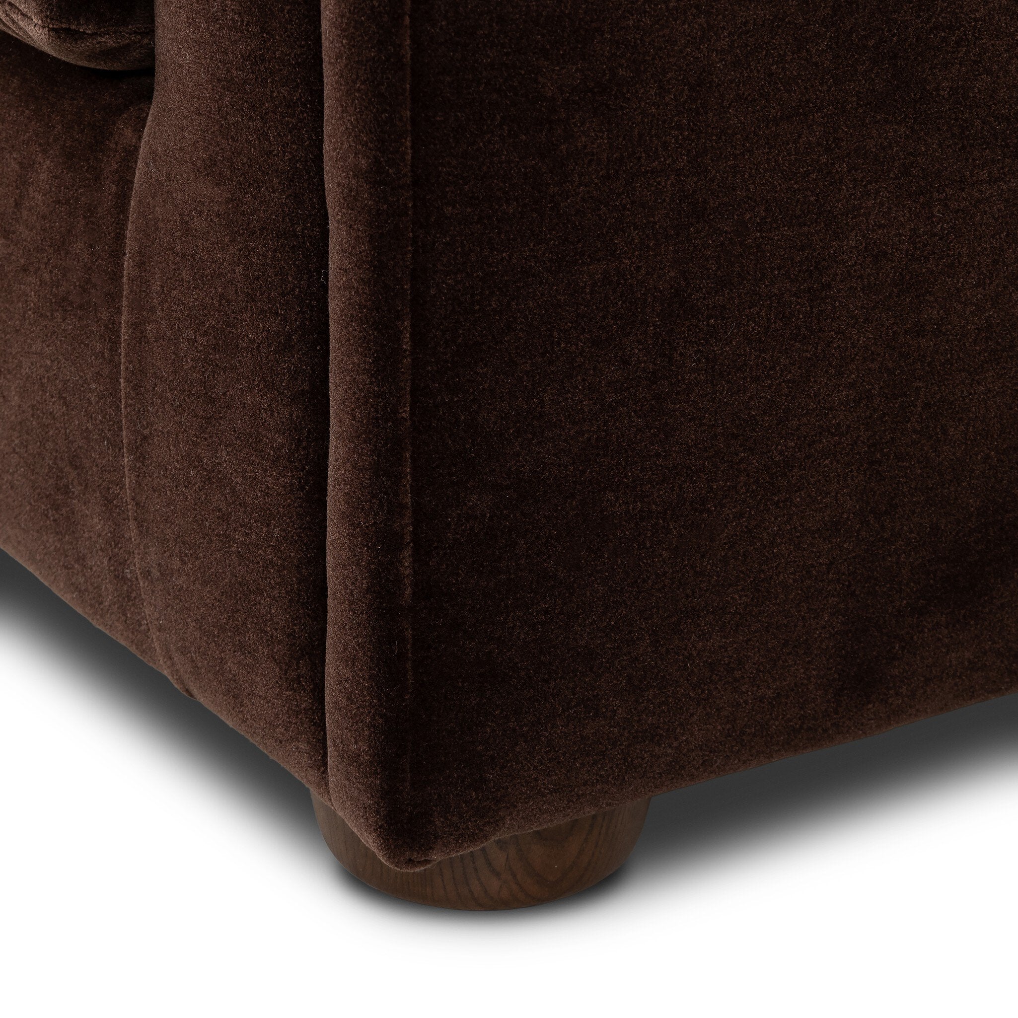 Close-up of a dark brown upholstered furniture corner showing the fabric texture and a small wooden foot visible at the bottom, on a white background.