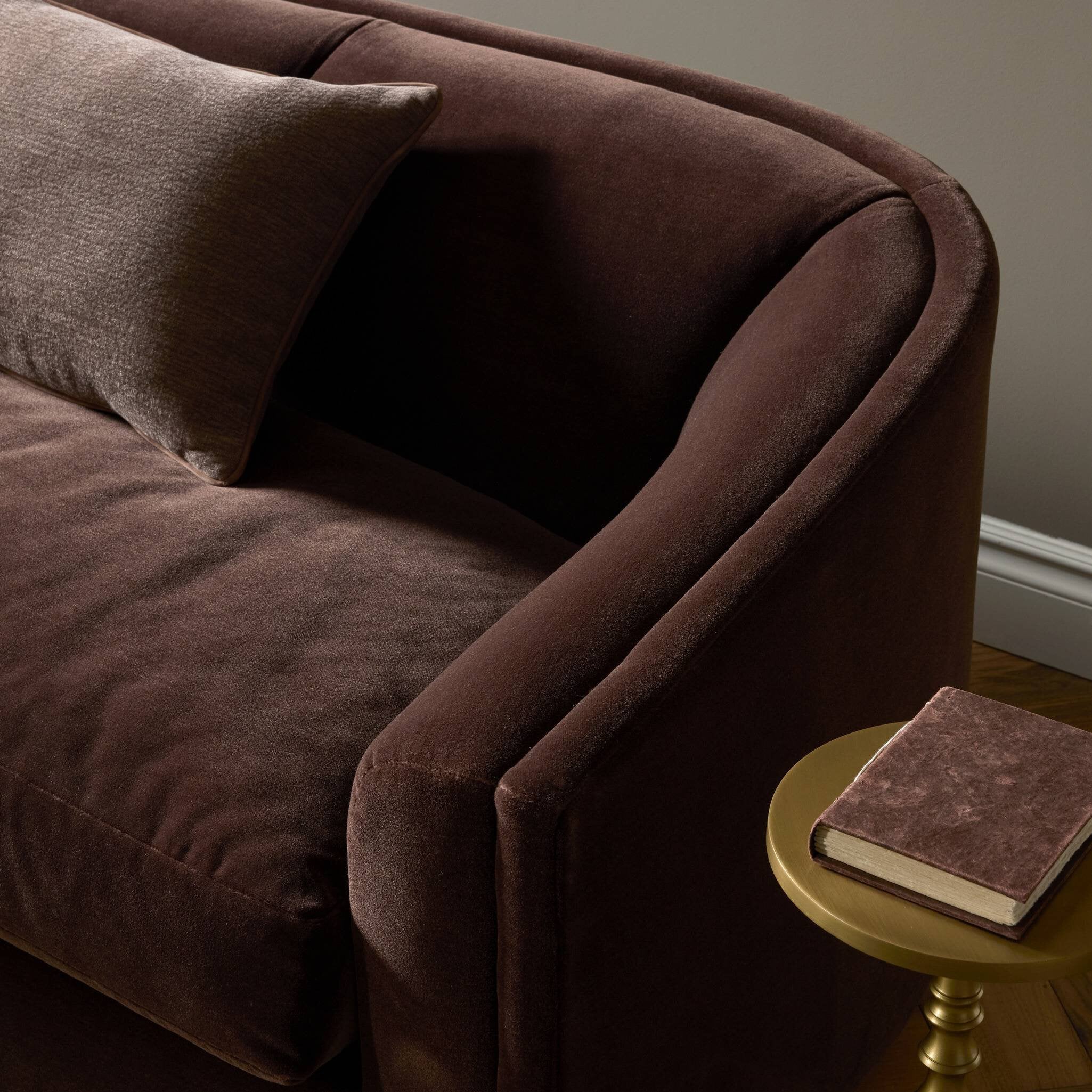 A close-up of a dark brown velvet armchair with a matching throw pillow, shown in a styled interior setting next to a small gold side table holding a book.