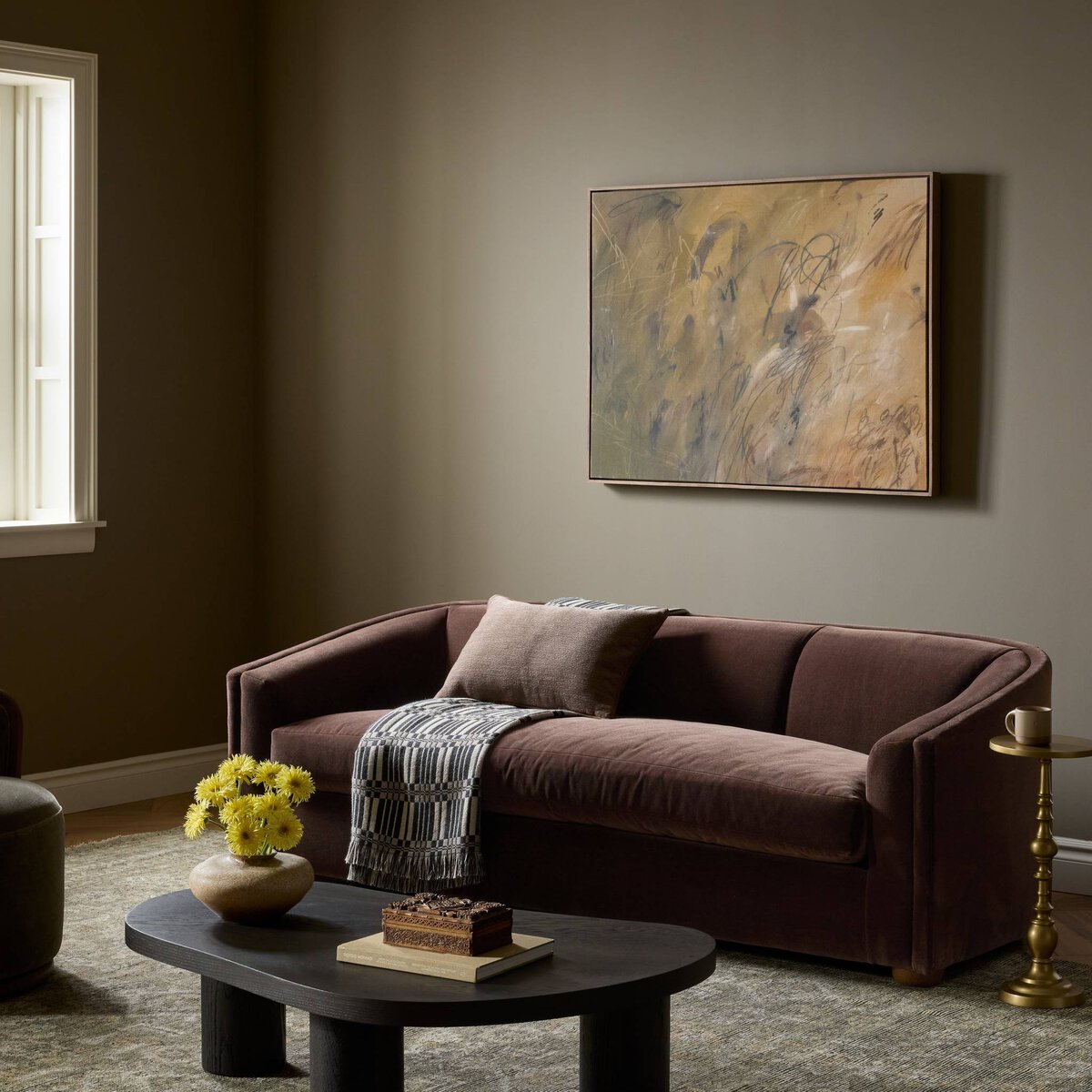 A styled living room scene featuring a dark brown velvet loveseat with a patterned throw blanket, set against a warm taupe wall with a large abstract framed painting. A dark wood coffee table with yellow flowers and a small side table are in the foreground.