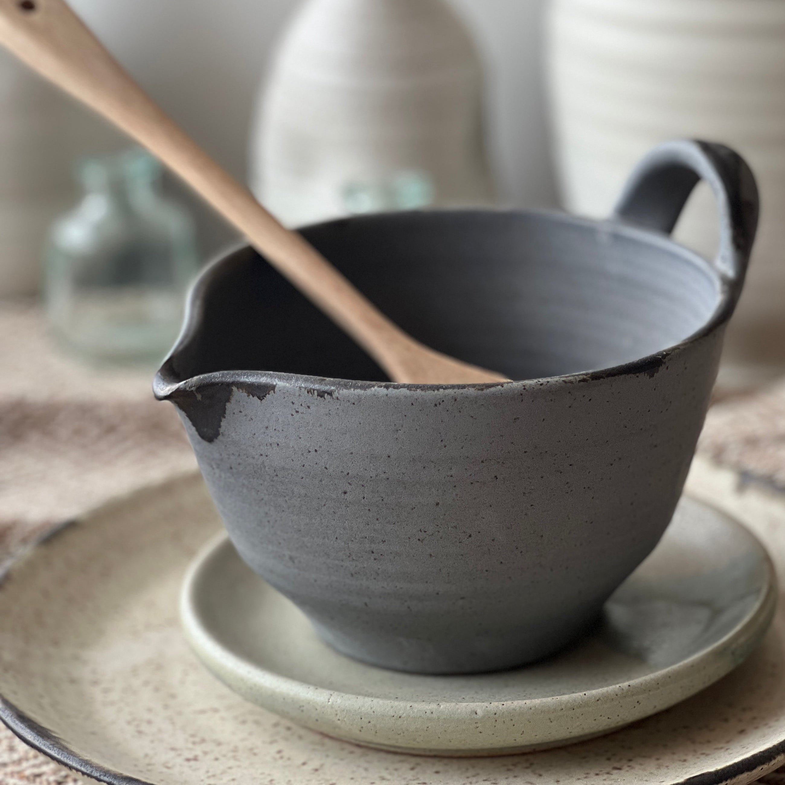 Gray ceramic bowl with wooden spoon on a textured surface with blurred ceramic vases in the background