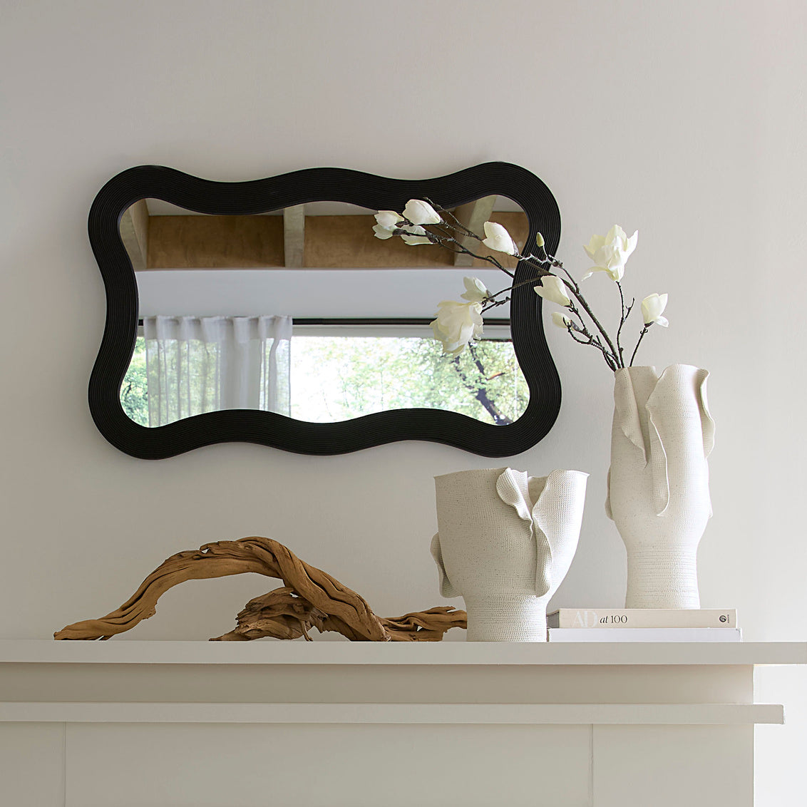 Decorative mirror with wavy frame on a white wall above a mantelpiece with vases and a piece of driftwood.