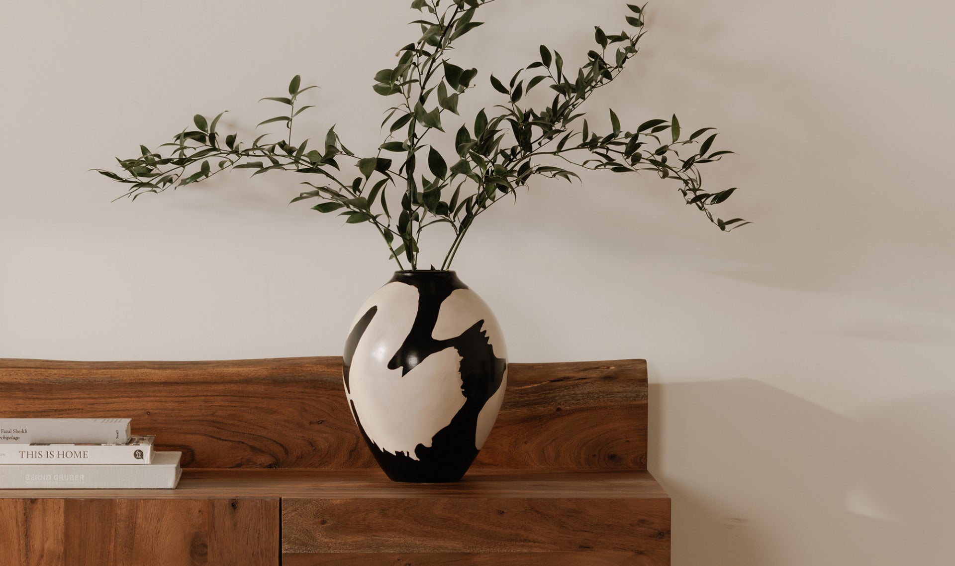 Black and white vase with green branches on a wooden sideboard against a plain wall