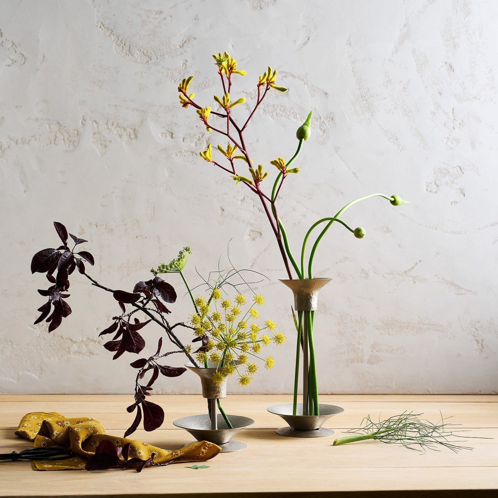 Three vases with floral arrangements on a wooden surface against a light gray wall.