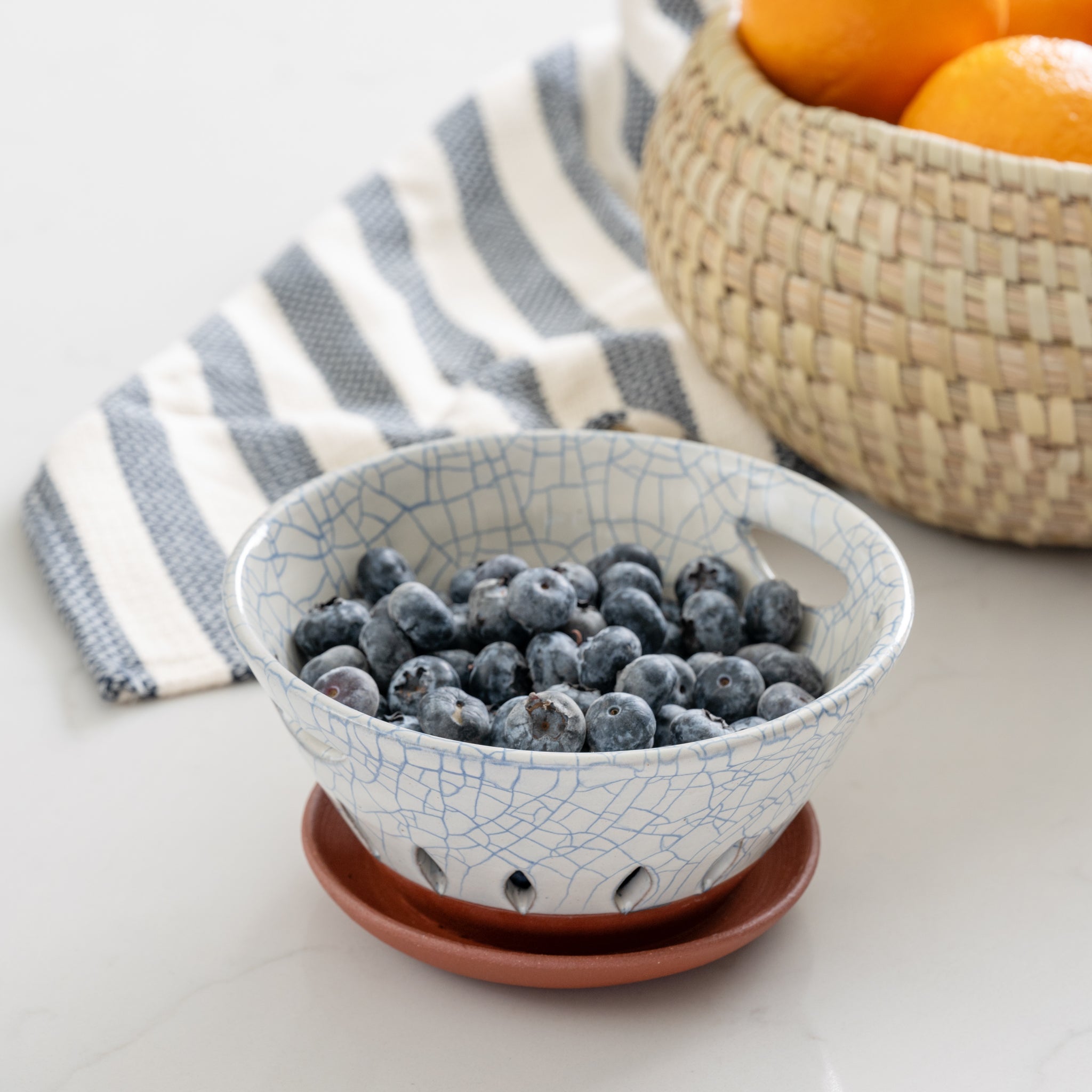 Ceramic berry bowl with blueberries on a white surface with a striped cloth and basket of oranges in the background.